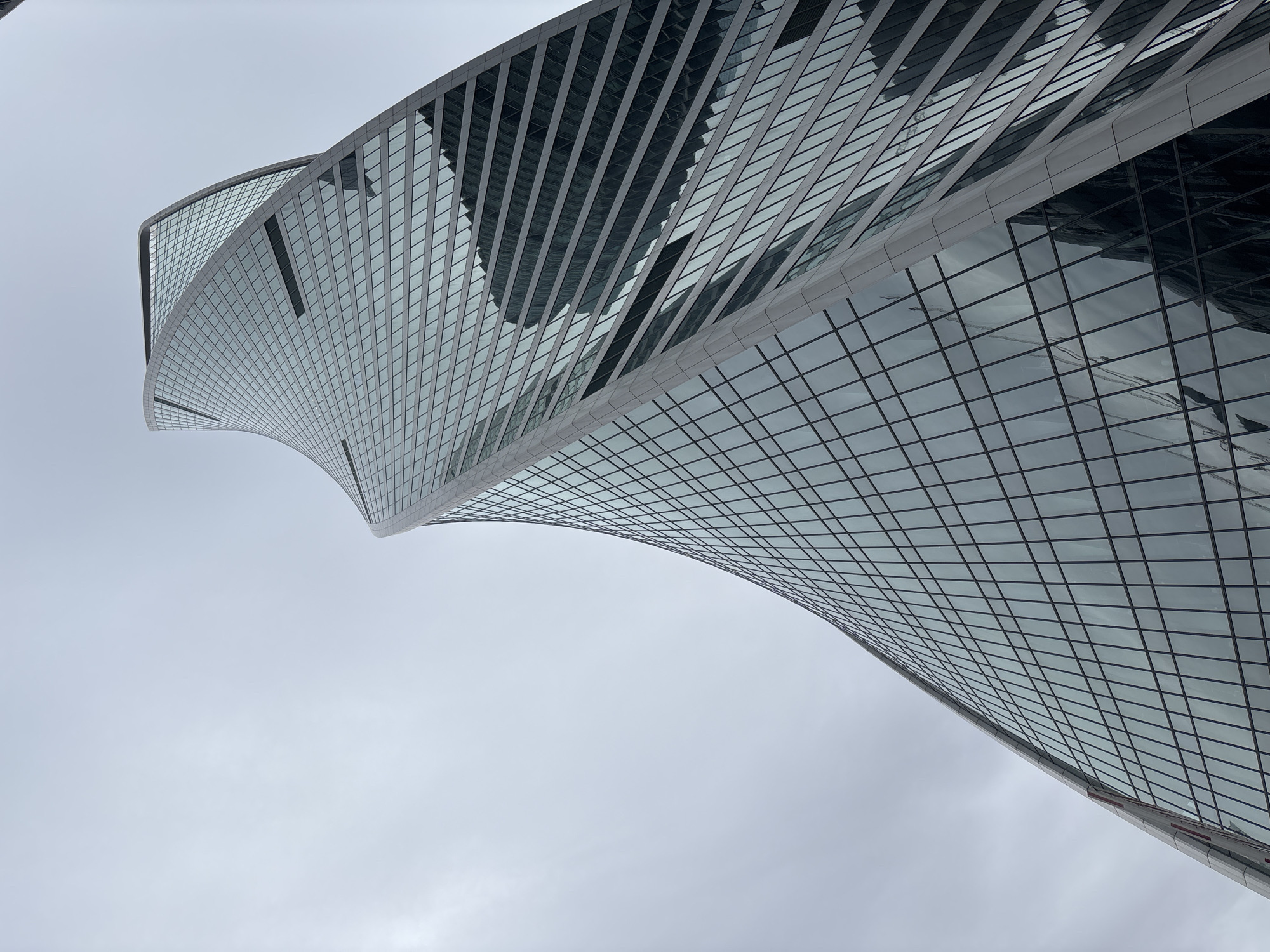 A low-angle, full shot depicts the Evolution Tower skyscraper in Moscow, Russia, against an overcast sky. The tower, known for its spiral shape, fills the upper portion of the frame. Its exterior is primarily glass with a grid pattern of dark lines, reflecting the surrounding buildings. The sky is a uniform gray, occupying most of the image area. The tower appears to be the sole focus, with no discernible people or other objects within the immediate vicinity.
