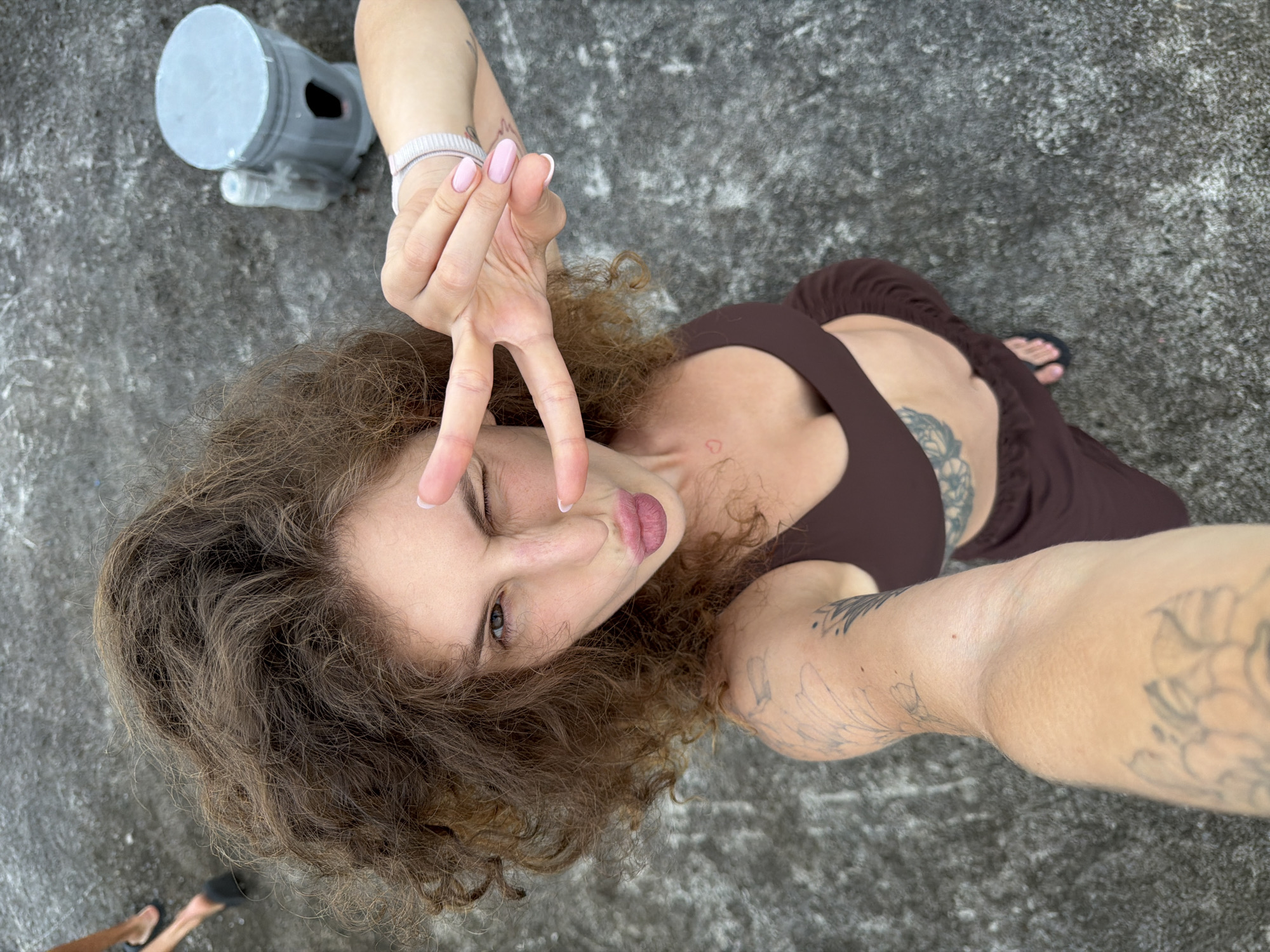A woman poses for a selfie. She stands on a gray concrete surface with some texture and discoloration. She wears a brown top and skirt that reveals a portion of her torso. Tattoos are visible on her chest, arms, and possibly back. She winks, makes a V sign with her fingers near her face, and pouts her lips. Her hair is curly and brown. A gray object resembling a piece of equipment with black inserts sits nearby. A foot clad in a black flip-flop appears in the bottom left corner. The location is likely North Kuta, Indonesia, based on available information.