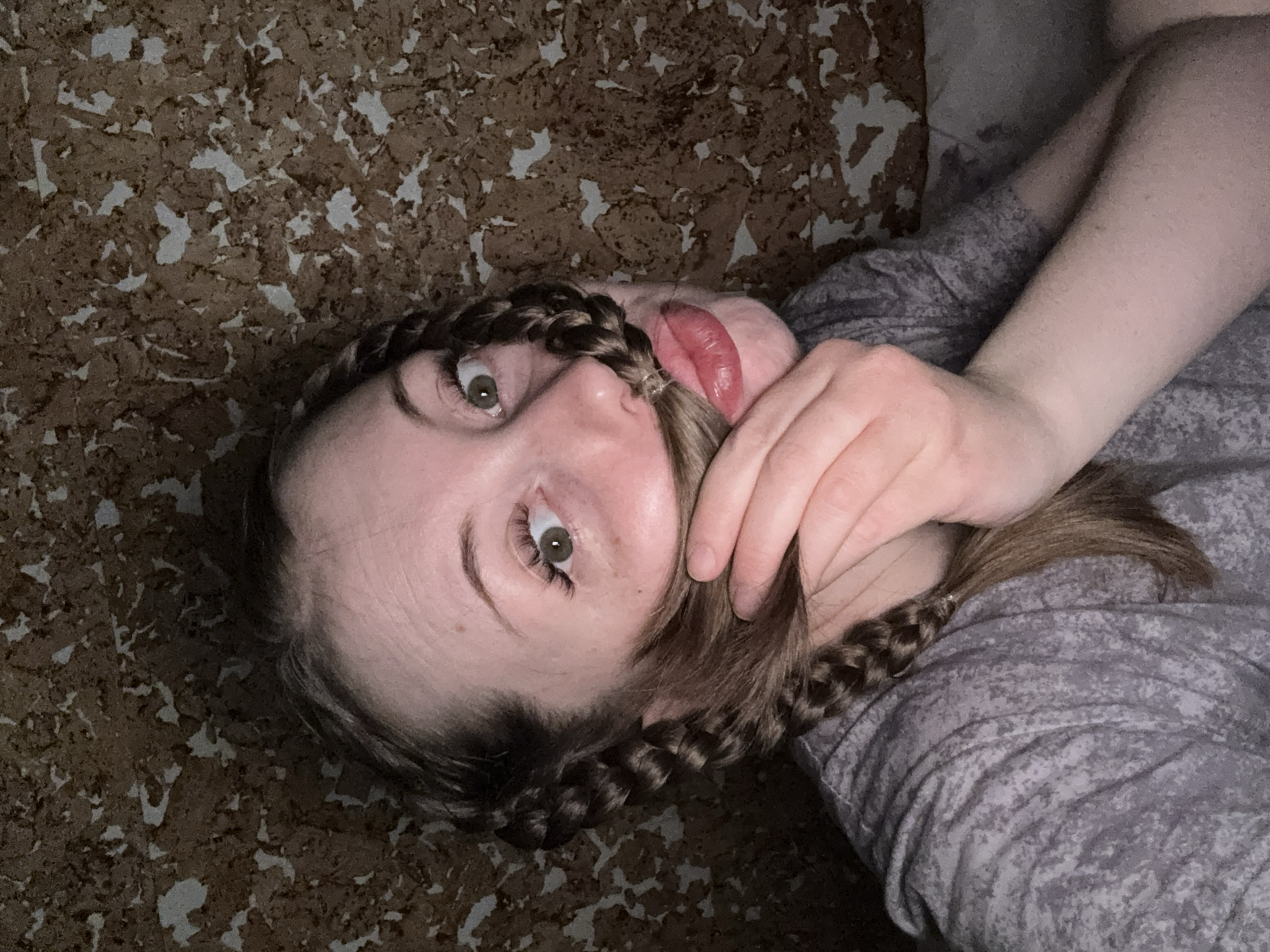 An overhead, slightly tilted shot captures a person lying down, looking upwards towards the camera. The subject has fair skin, prominent green/hazel eyes, and dark brown hair styled in two braids, one visible on each side of their head. One hand is raised, gently holding a strand of hair, which appears to be playfully held in their slightly open, red-tinted lips. The person's expression is one of playful curiosity or engagement, with their eyes wide open. They are wearing a grey, long-sleeved garment with a subtle mottled or patterned texture. The background is a textured, mottled brown surface with irregular lighter beige or white patches, resembling a cork board or patterned wallpaper/flooring. No text is visible in the image. The lighting suggests an indoor setting.