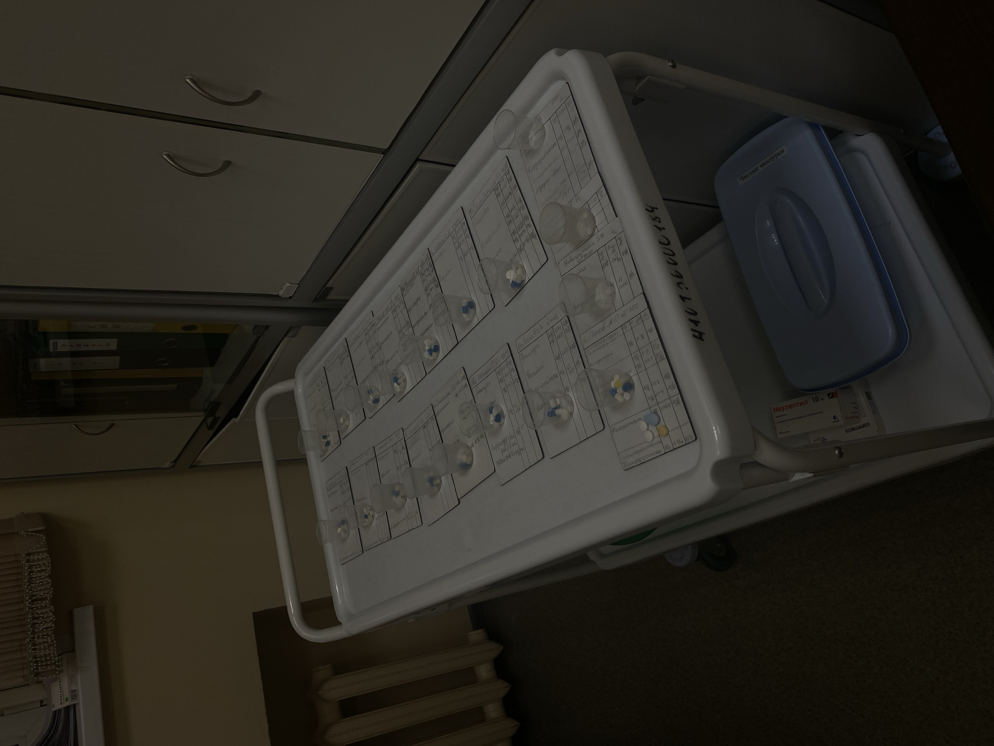 The image depicts a medication cart in what appears to be a medical facility, possibly a hospital or clinic, in Saint Petersburg, Russia. The cart, made of white metal, is laden with individual cups containing pills and capsules, arranged on sheets of paper. These sheets are organized into rows and columns, suggesting a system for dispensing and tracking medications for patients. Some of the pills are white, while others are blue. The cart has a white handle and is on wheels. In the background, a radiator and a wall with cabinetry are visible. On the lower shelf of the cart, there is a blue medical box with a handle and Russian text that reads "Чистые Маршруты" (Clean Routes). The lighting is dim, casting shadows and contributing to a sterile, functional atmosphere. The overall scene is consistent with a medical setting where medications are prepared for distribution.