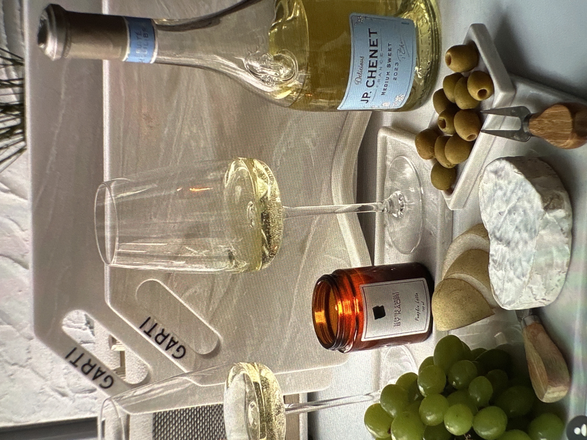 The image presents a still life arrangement featuring culinary and decorative elements, likely staged for a photograph. Dominating the scene is a bottle of J.P. Chenet medium sweet white wine from France, vintage 2023, placed alongside two partially filled wine glasses. Accompanying the wine is a curated assortment of food items, including green olives, a segment of brie cheese, pear slices, and a bunch of green grapes. A small cheese knife rests near the brie and pear. An amber-colored scented candle labeled "Scented Candle By Kaori, Pumpkin Latte" adds to the ambiance. The entire arrangement is placed on a light-colored serving tray branded with the name "GARTI". The background appears to be a textured wall. The image provides no information about people or location in the city of Yerevan, Armenia.
