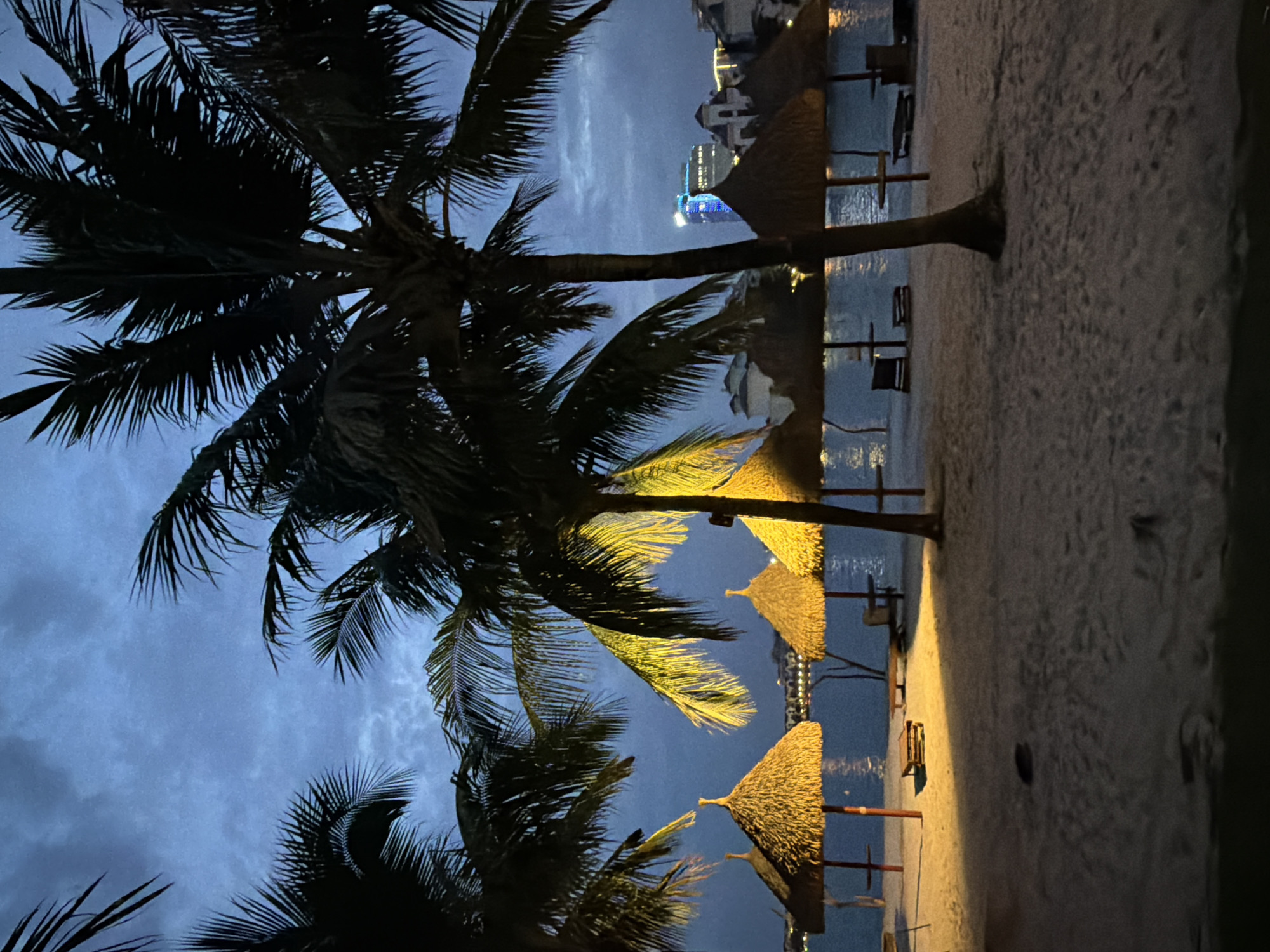 The image shows a beach at dusk in an urban waterfront area of Hanoi, Vietnam. Several thatched umbrella structures are arranged on the sandy beach, some of which are illuminated by artificial light. Palm trees with dark fronds are silhouetted against a twilight sky. In the background, across a body of water, there are buildings and lights indicating a city skyline. The water appears calm, reflecting the ambient light. There are no people visible in the scene. The overall atmosphere is tranquil and nocturnal.