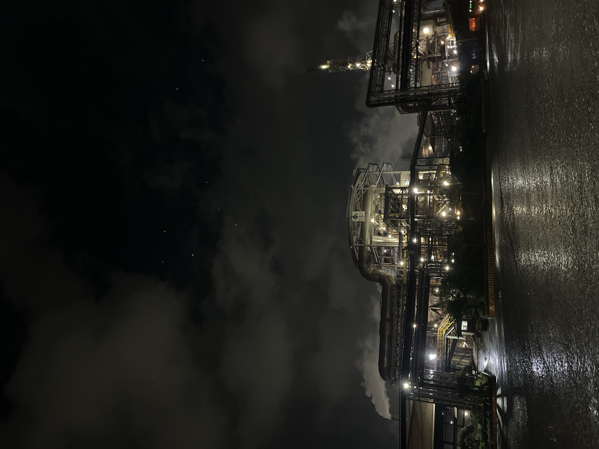 The image depicts an industrial facility at night. The location is Unknown, Indonesia. The facility is comprised of multiple interconnected structures of steel and pipework illuminated by artificial lighting. The sky is overcast with clouds. A wet surface, likely a road, occupies the foreground. There is no visible human activity.