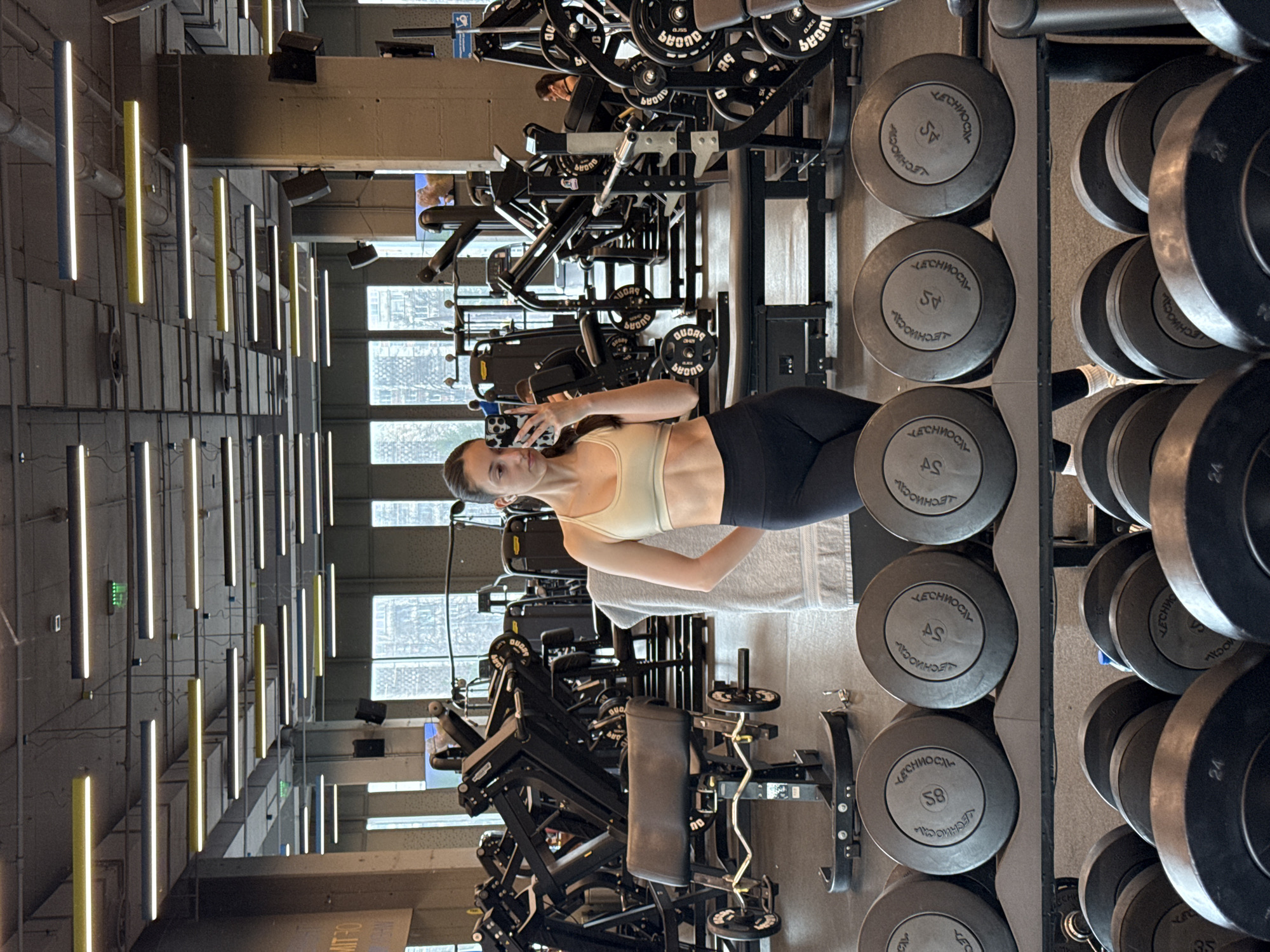 The image depicts a female individual taking a mirror selfie inside a modern fitness center, identified as Pulse Fitness & Spa MITRO in Sofia, Bulgaria, based on visible branding in the background. The subject, a young adult female with dark hair pulled back, stands centrally, clad in a light-colored sports bra and black athletic leggings, holding a smartphone with a black and white patterned case in her right hand. A white earbud is visible in her left ear.
In the foreground, a black, multi-tiered rack holds numerous black dumbbells, each featuring "ЛЕЧНОСТИ" (LECHNOSTI) and numerical weights (e.g., 58, 45, 24) embossed in white Cyrillic lettering. Directly behind the subject, a light gray towel is draped over a piece of gym equipment. The expansive midground and background contain a variety of black Technogym weight machines and benches, including an arm curl bench with a barbell. Large, multi-pane windows line the far wall, providing a view of exterior urban buildings and a distant spire-like architectural feature.
The facility features an exposed concrete ceiling from which numerous linear LED light fixtures, alternating in yellow, gray, and white, are suspended. Visible ventilation ducts and structural elements are also present. Additional gym patrons are partially visible operating equipment in the background. The floor is a dark, likely rubberized, gym surface. Wall-mounted speakers and display screens are also evident within the gymnasium environment.