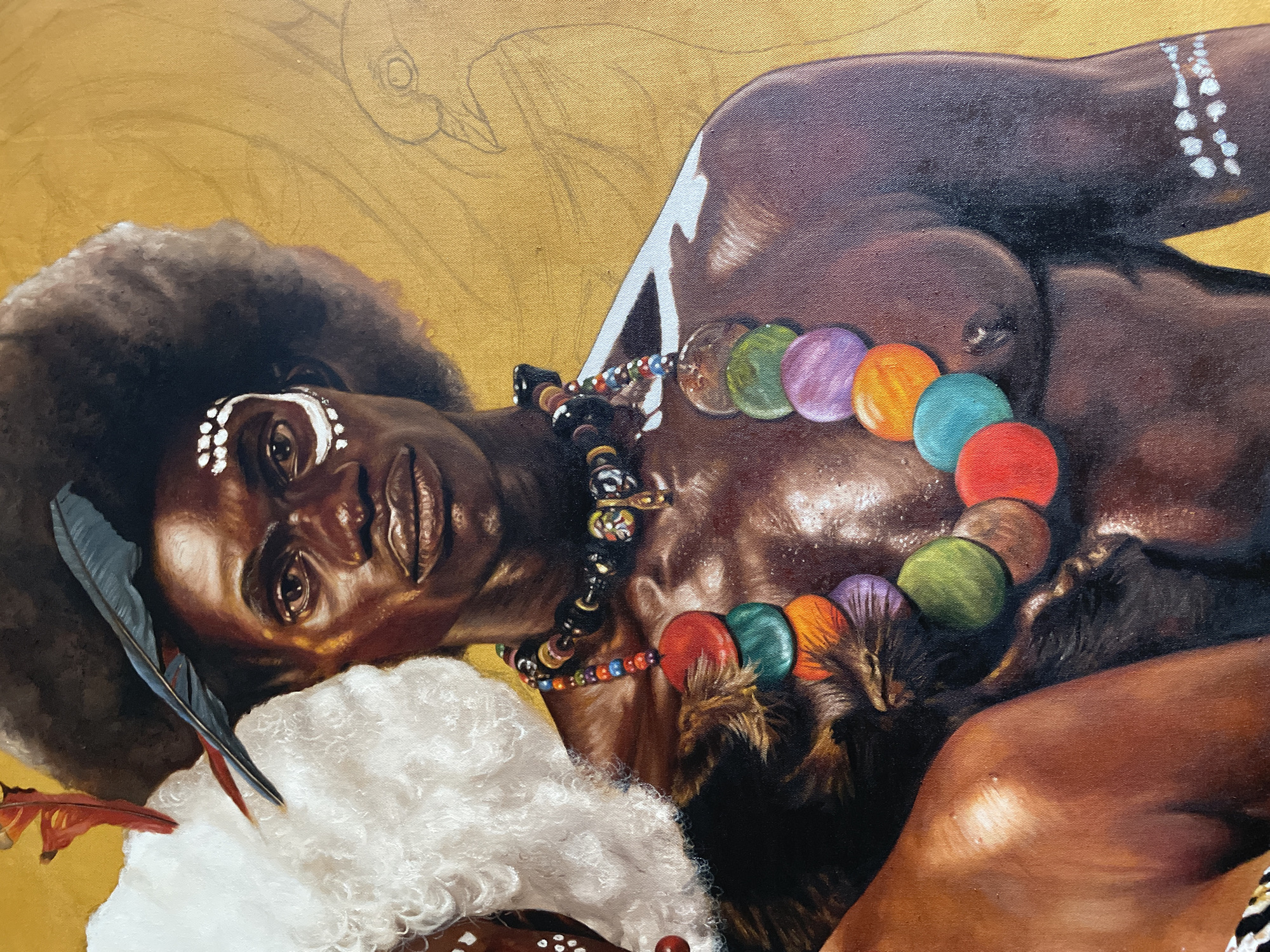 The image shows a painting of a dark-skinned person lying down. The person has a large afro and is wearing a colorful necklace made of large circular beads and other beaded necklaces. There are white markings on their face, and they are lying on a fluffy white surface. Feathers are placed near their head. The background is a solid golden color with faint outlines of a drawing visible in the top left corner. There is no visible location in the city of Ado, Nigeria in the image.