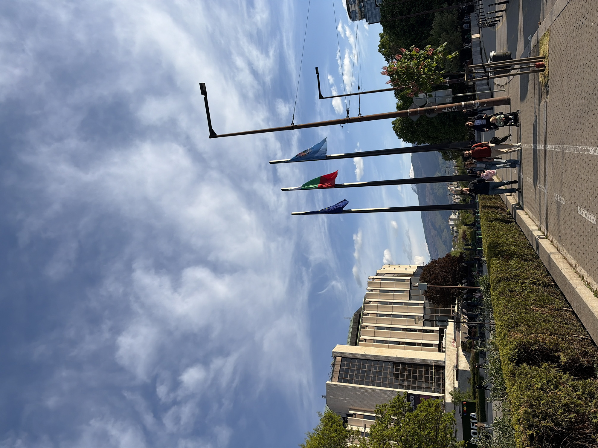 The image depicts a street scene in Sofia, Bulgaria. Three flags hang from poles. From top to bottom, they appear to be an unidentified light blue flag, the Bulgarian flag (white, green, red), and another unidentified dark blue flag. A group of pedestrians, including a family with a child in a stroller, are walking along a sidewalk paved with gray rectangular blocks. The sidewalk is bordered by a trimmed hedge. A multi-story building is to the left of the hedge, with trees in front of it. Mountains are visible in the background. The sky is blue with scattered white clouds.