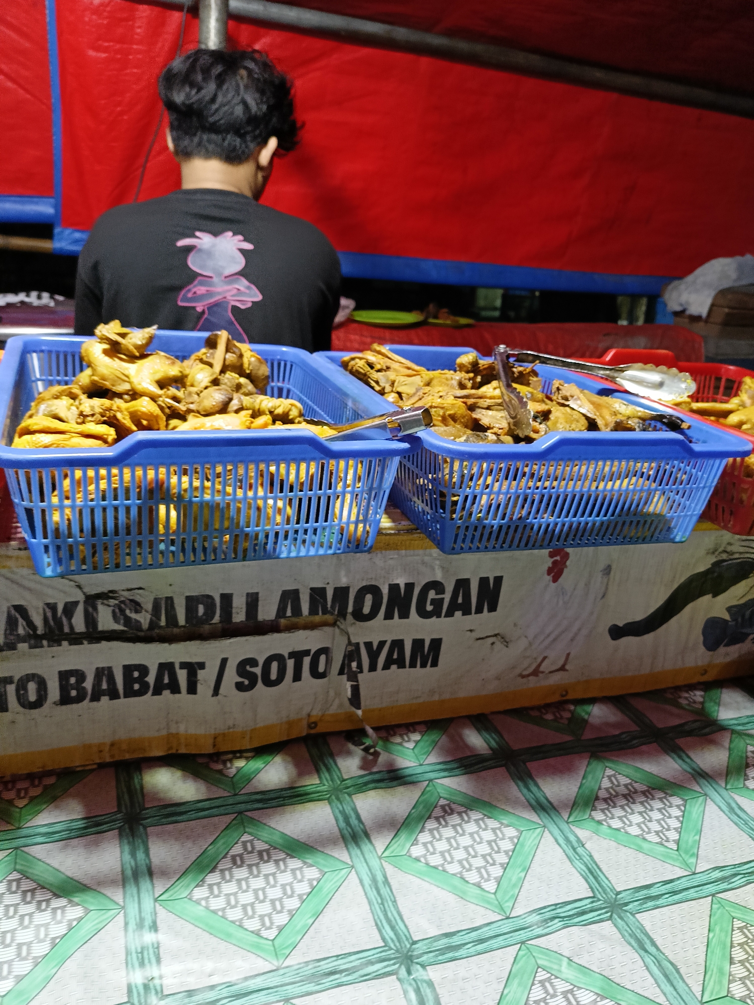 The image shows a street food stall in Kebondalem Kidul, Indonesia. A person wearing a black t-shirt with a pink graphic is standing behind two blue plastic baskets filled with fried chicken. The stall has a red backdrop and a sign that reads "TAKI ADILI LAMONGAN" and "TO BABAT / SOTO AYAM". The foreground shows a patterned green and white tablecloth.