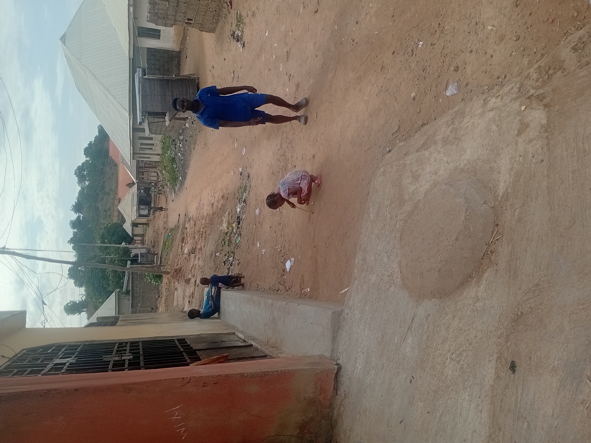 The image shows an outdoor scene in Unknown, Unknown. Three people are visible. An adult male, wearing a blue shirt and shorts, stands in a dirt road. A child in a light-colored dress is crouched, seemingly playing in the dirt. Another person is sitting along a concrete structure that borders the road. Buildings and trees are visible in the background. The ground appears to be dirt and concrete. There is litter scattered around.