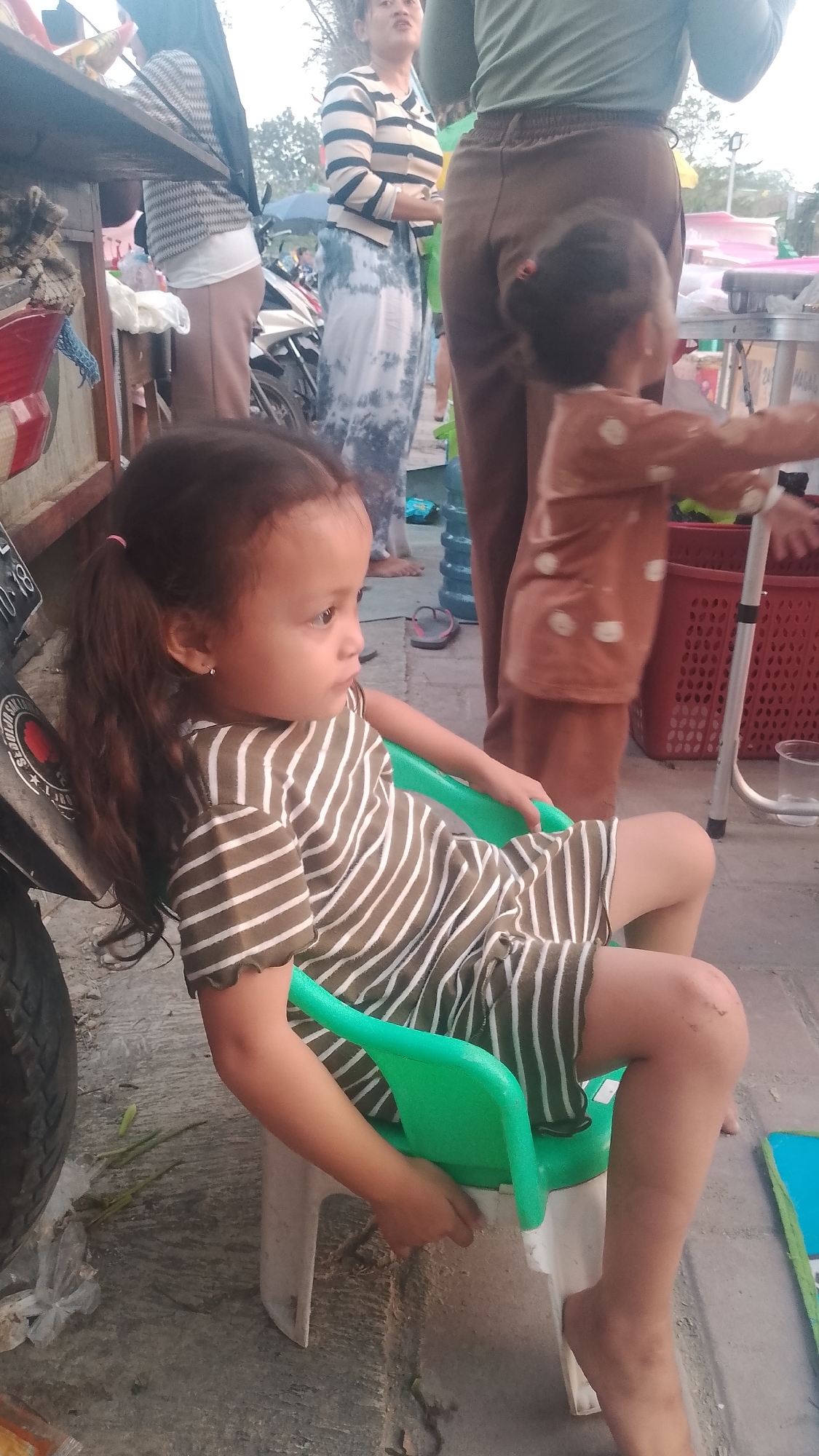 A young girl with dark hair pulled into two pigtails, wearing a green and white striped dress, sits on a green plastic chair. Her bare feet rest on the ground. She is looking to her right. Behind her, a person in brown pants and a green shirt is standing. Another person in a black hijab and a patterned top, and a person in a striped shirt and a white and blue tie-dye skirt are also visible in the background. The scene appears to be outdoors, possibly a street or market, with a motorcycle and a cart visible. The ground is paved with concrete. There are blue water jugs and red plastic baskets in the background. The location is Karanglangu, Indonesia.