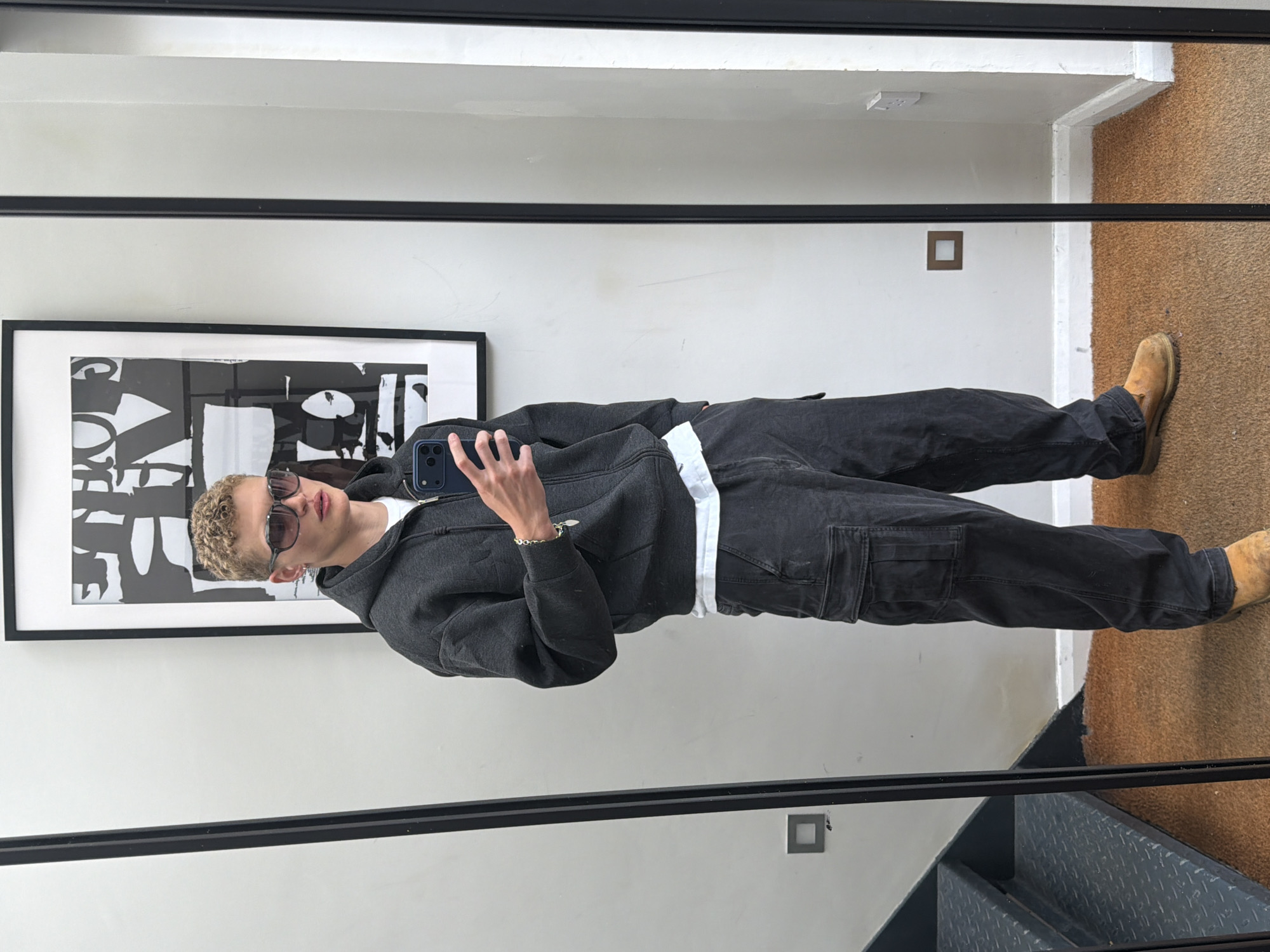 This mirror selfie, taken in London, United Kingdom, captures a young individual standing reflected in a large mirror. The person, who has short, curly, light-colored hair, is wearing dark sunglasses and holding a dark blue smartphone in their right hand, angled to take the picture.
They are dressed in a casual outfit consisting of a charcoal or faded black full-zip hooded sweatshirt, worn over a white t-shirt that is visible at the hem. Matching dark grey or faded black cargo pants cover their legs, and they wear sturdy light brown or tan work boots. A subtle gold-colored chain bracelet adorns their right wrist.
The setting appears to be a modern indoor space. Behind the person's reflection, a white wall is visible, on which a large, framed abstract black and white artwork hangs. This artwork features bold, graphic, letter-like shapes, contributing to a contemporary aesthetic. The floor in the reflection is covered with a light brown, possibly carpeted, material that extends to the right. A prominent black metal staircase with diamond-plate treads, along with a matching handrail, is visible towards the bottom right, suggesting the image was taken near a stairwell. A small, dark square object, possibly a light switch or outlet, is mounted on the white wall. The lighting throughout the scene is even, indicative of an indoor environment, likely during daytime. No specific legible text is visible other than the abstract shapes within the artwork.