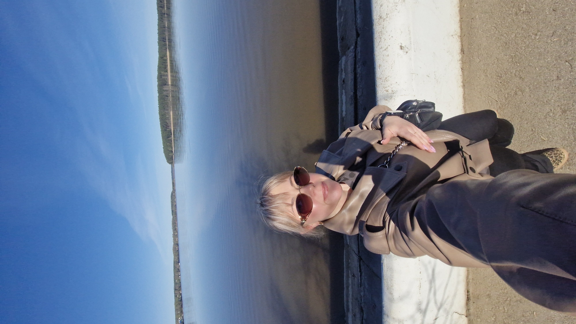 A medium, eye-level shot shows a woman seated next to a body of water on a sunny day in Otchor, Russia.
The woman, positioned to the right of the frame, wears a light brown leather jacket, black pants, and leopard-print shoes. She also has on sunglasses and a black purse hanging from her shoulder. Her nails are painted with pink nail polish. She sits on a gray concrete surface bordered by a white wall.
To the left of the woman, a large body of water stretches across the frame. A narrow strip of land with trees lines the opposite shore. A clear blue sky is visible above the water. The scene is well-lit, and the shadows are distinct.