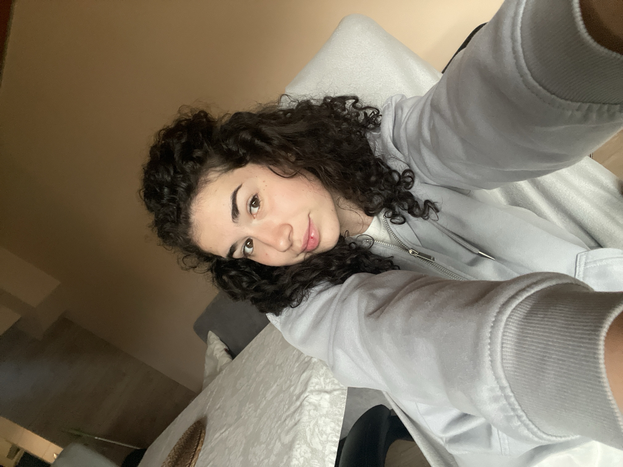 A young person, likely female, is captured in a close-up, angled selfie. She has dark, voluminous curly hair that frames her face, and warm brown eyes. Her expression is calm and inviting, with a subtle, gentle smile directed towards the camera. She is wearing a light grey zippered hoodie over a white top, with the textured fabric of the hoodie's sleeve prominent in the lower right foreground.
The setting appears to be indoors, likely a domestic environment such as a living room or bedroom. A soft, white pillow or cushion is visible behind her head, and a light-colored, possibly patterned tablecloth or blanket covers a surface beneath her arm. A small, woven basket can be seen on this surface. The background consists of a plain, light brown or beige wall. The lighting suggests it is daytime, providing a soft illumination without harsh shadows. No visible text is present in the image. This indoor scene is situated in what is understood to be Sofia, Bulgaria.