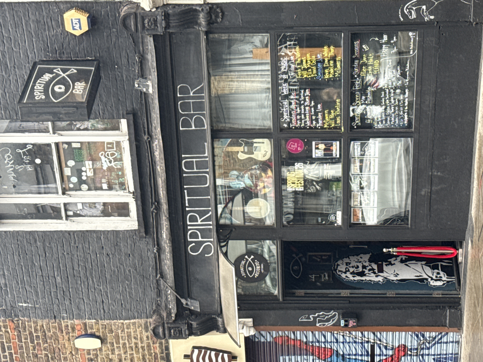 The image shows the exterior of "Spiritual Bar" in London, United Kingdom. The building is painted black, with a sign reading "Spiritual Bar" above the entrance. A smaller sign with a fish symbol also reads "Spiritual Bar". The windows display a schedule of musical performances. The door has a mural on it and a red rope barrier. There's an ADT security sign on the upper-left wall. A window is located to the left of the bar entrance. The storefront is adorned with graffiti.