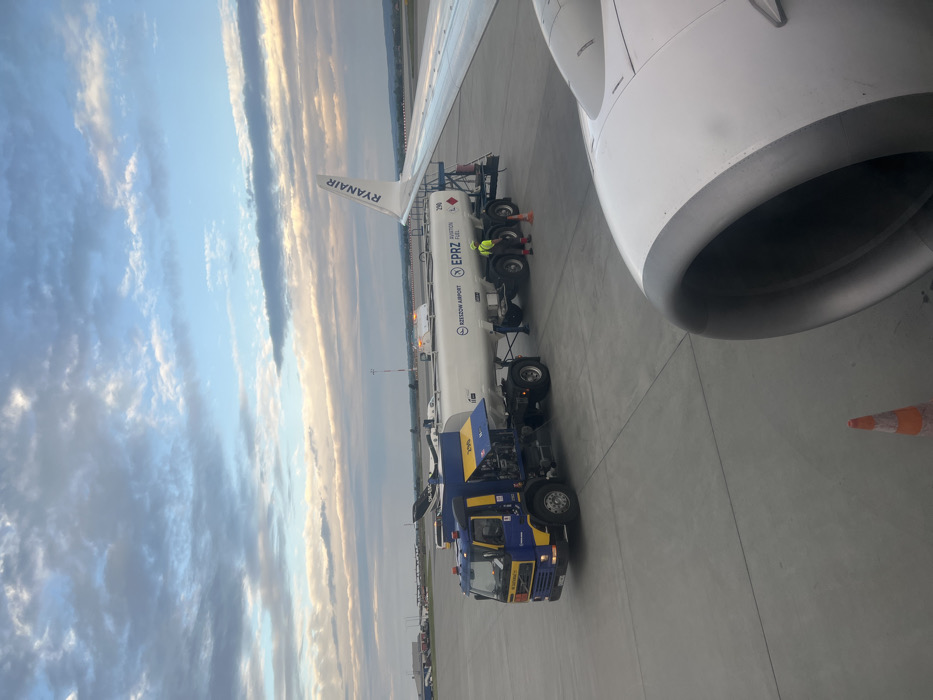 The image shows a fuel tanker truck connected to a Ryanair airplane, refuelling it. The airplane is parked on a tarmac at Rzeszow Airport, located in the city of Jasionka, Poland. The coordinates provided are (latitude: 50.114, longitude: 22.026). The sky is mostly clear, with some white clouds. There is a lone person standing beside the truck, and the airplane is partially visible in the foreground. There are other airplanes in the background. The ground is paved. A bright orange traffic cone is visible in the bottom right of the frame. The fuel truck is labelled "Rzeszow Airport" and "EPRZ Aviation Fuel".