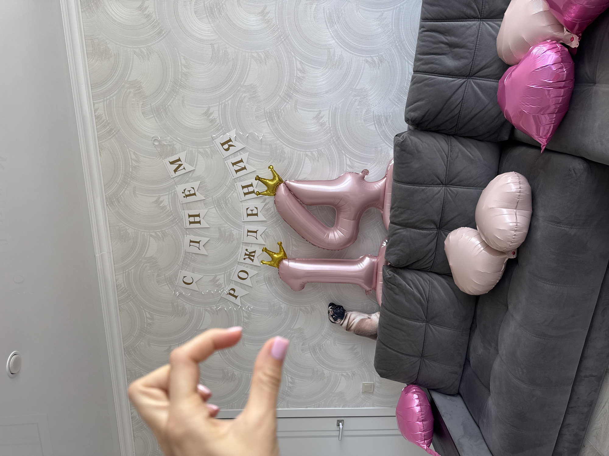The image is an indoor shot, likely taken in a residential space, and it does not directly contain enough information to ascertain that it was taken in Sochi, Russia. The scene includes decorations for a birthday celebration, specifically the number 14 rendered in pink balloons with small golden crowns attached. A banner spelling out "С днем рождения" (Happy Birthday) is affixed to the wall above the balloons.
A gray sofa is present on the right side of the image, adorned with several pink and light pink heart-shaped balloons. Part of a person's hand, with nails painted light pink, is visible in the lower-left corner, suggesting someone is taking the photograph. A pug is partially visible in the balloons with crown.
The backdrop consists of patterned wallpaper in a neutral color. The photograph is rotated. A door and door knob are partially visible in the bottom left.