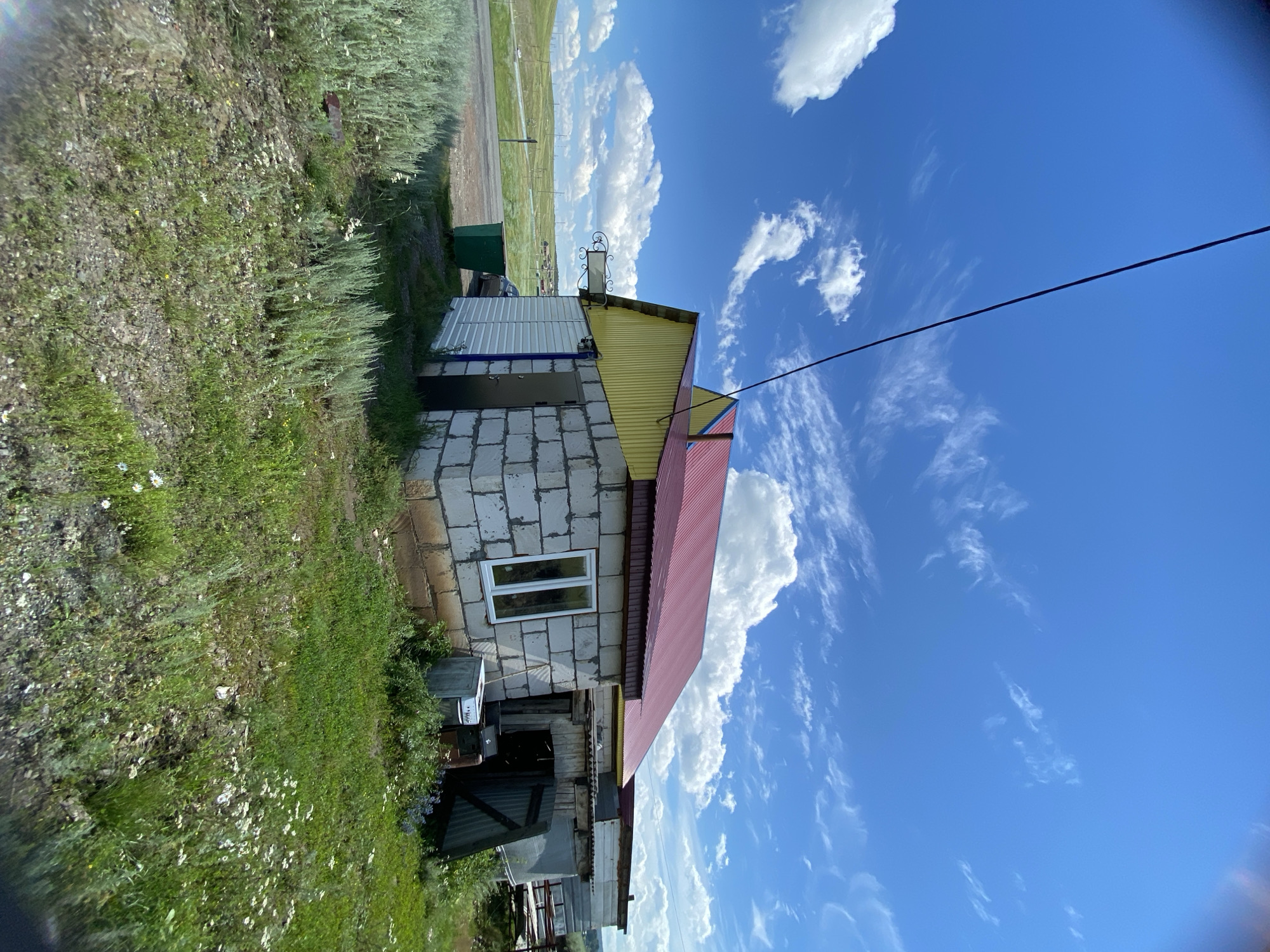 The image depicts an exterior view of a building complex in Yuldybayevo, Russia. The primary structure is a single-story building with a red metal roof and yellow siding. It is constructed from white cinder blocks and features a white-framed window and a dark metal door. Adjacent to this building is an attached structure with a corrugated metal roof. The scene is set against a backdrop of a bright blue sky with scattered white clouds. A power line stretches diagonally across the upper portion of the frame. The immediate surroundings are characterized by a grassy area with sparse wildflowers, some scrub brush, and a green trash receptacle. In the background, a road is visible, with a car parked near the building. The landscape beyond appears to be rolling hills covered in vegetation.