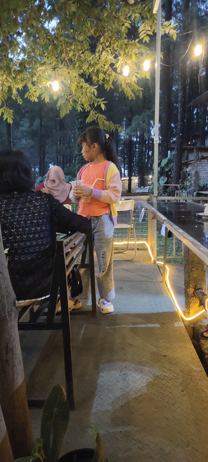 An outdoor evening scene in Cembor, Indonesia, features three individuals on an elevated platform illuminated by string lights. In the foreground, facing away from the camera, a person with dark hair wears a black patterned top, seated on a dark metal bench. To their right, a young female, approximately 8-12 years old, stands facing left, looking into the distance. She wears an orange T-shirt, a multi-colored striped cardigan (yellow, pink, purple), light blue ripped jeans, and white Crocs. Her dark hair is pulled back into a ponytail with a yellow tie. She holds a clear plastic cup with a straw in both hands. Behind the seated person, partially visible, another individual wears a light brown hijab.

The setting appears to be an outdoor cafe or restaurant with a rustic aesthetic. The ground is a dark metal checker plate material, bordered by a light-colored railing on the right, decorated with a yellow LED strip light. A dark, possibly wet, tabletop is visible beyond the railing. In the background, numerous tall, dark trees, resembling pines, indicate a wooded environment, suggesting dusk or nighttime. Overhead, a tree canopy with green leaves is visible, from which string lights with exposed bulbs hang, providing warm illumination. Scattered around are various outdoor furniture pieces, including white chairs, and potted plants. A small, multi-level structure with stone or brick elements is discernible in the far right background amidst the trees.