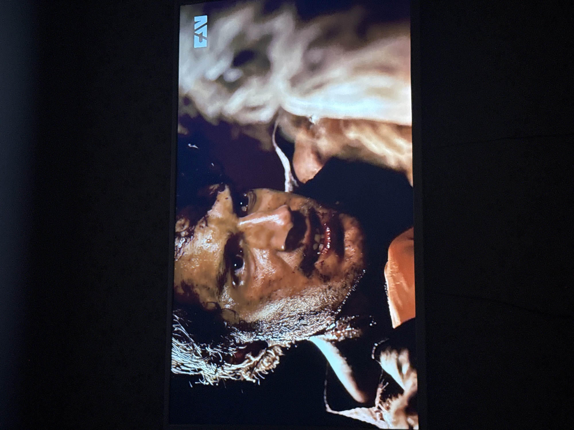 A medium close-up shot depicts a man's face, appearing disheveled and possibly injured, as the image is dark and somewhat indistinct. The man has dark hair and a beard, and his face seems to be covered in dirt or grime. Overlapping the image of the man is a blurred image of a woman with blonde hair. The image is being displayed on a screen, indicated by the presence of the logo "FX" in the top left corner. There are no indicators within the image to suggest the location as Mytishchi, Russia, or any other specific location.