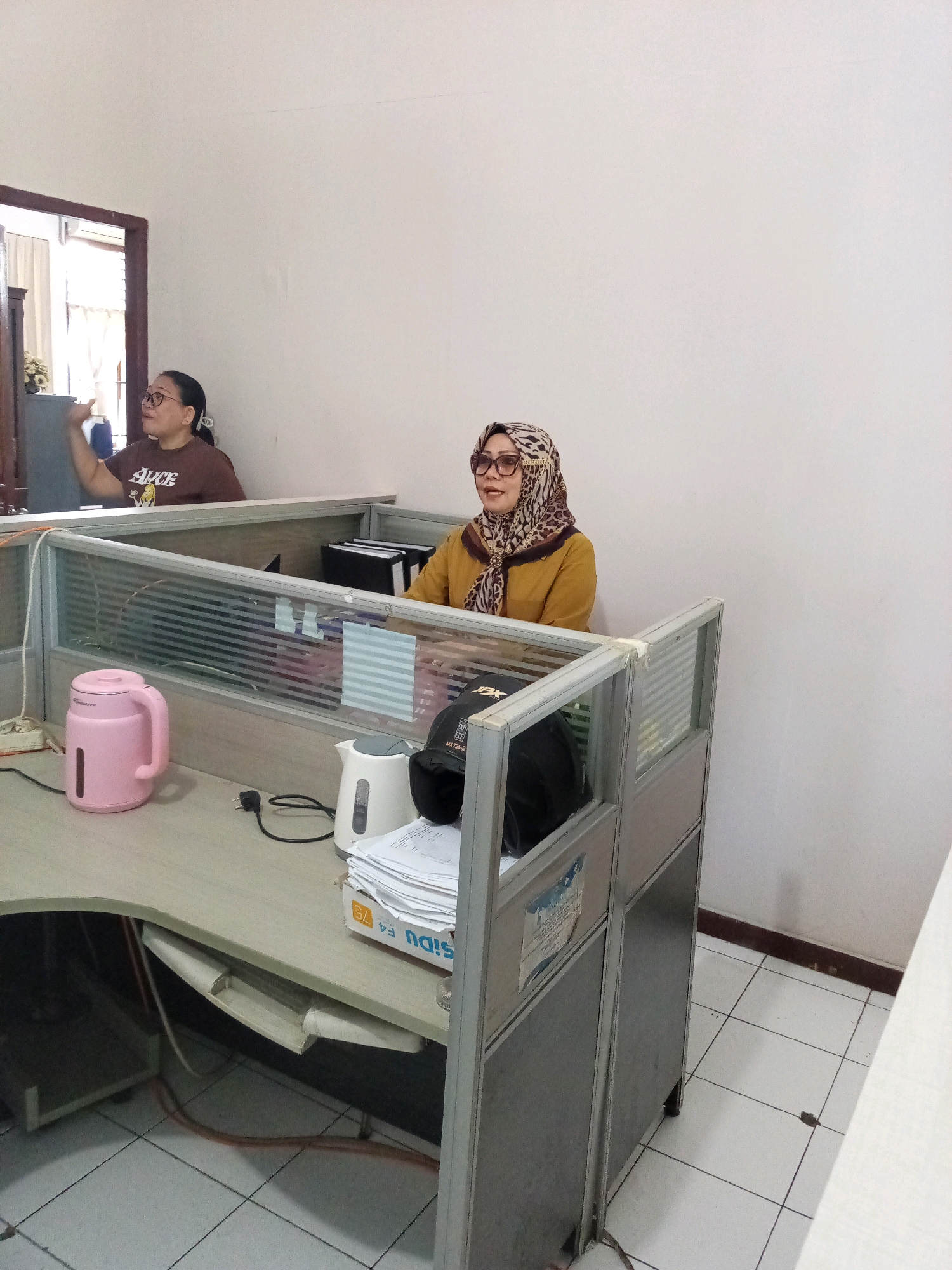 The image depicts an indoor scene, likely an office environment, with two women present. One woman, wearing glasses and a brown t-shirt with the word "ALICE" printed on it, is seated at a desk to the left. She is gesturing with her left hand. The second woman, wearing a leopard-print hijab and an orange top, is seated at a desk to the right. She is looking towards the left. Both women appear to be working at their respective desks, which are separated by cubicle partitions. On the desk to the left, there is a pink electric kettle. On the desk to the right, there is a white electric kettle and a stack of papers. The floor is tiled. The location is identified as Bitung, Indonesia.