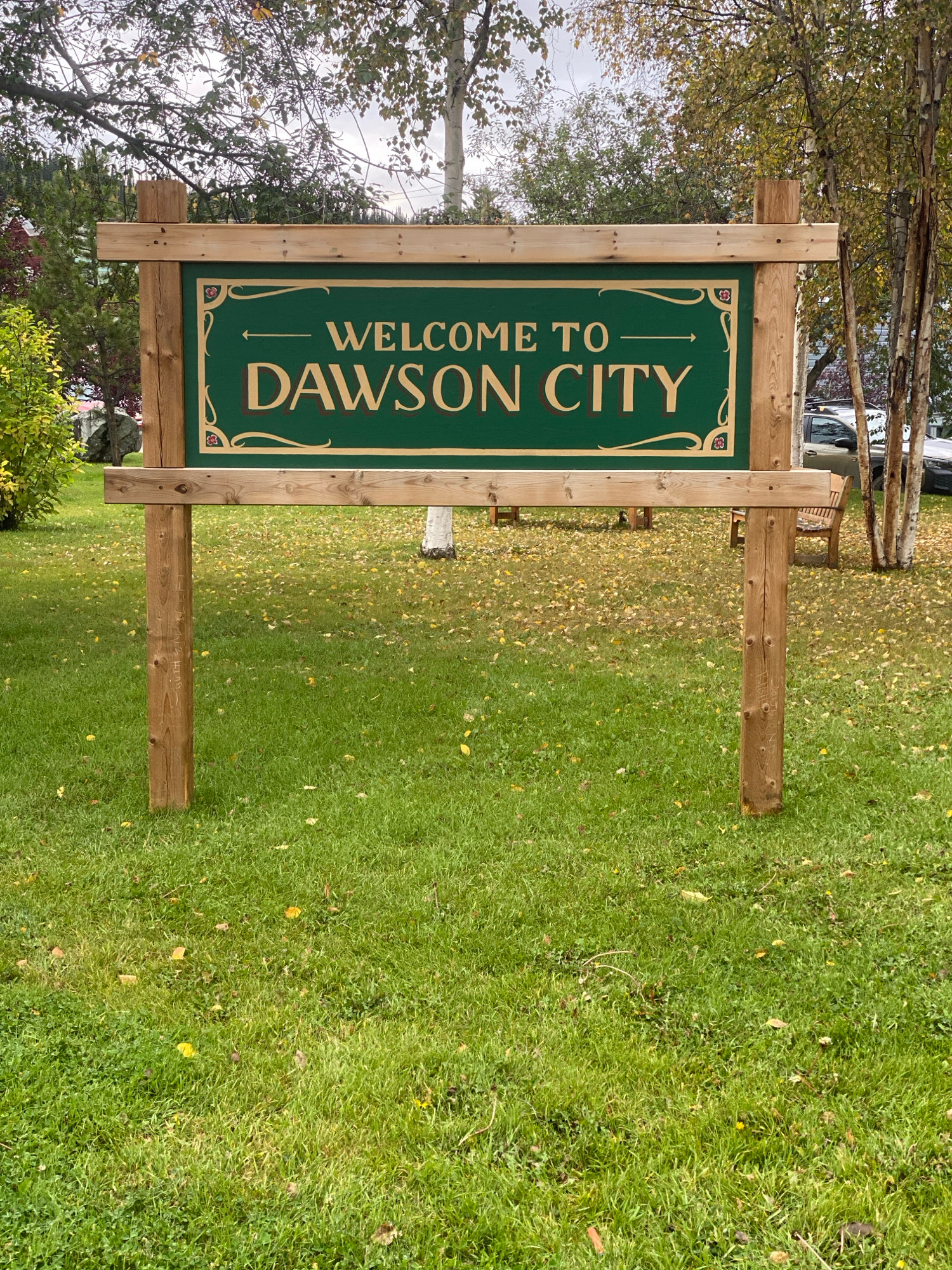 A sign with "WELCOME TO DAWSON CITY" in large, decorative lettering welcomes visitors. The sign is green with a gold border and has ornate flourishes. It is supported by four wooden posts, two in front and two in back, constructed from rough-hewn lumber. The sign is set in a grassy area dotted with fallen leaves, suggesting an autumn setting. In the background, trees with autumnal foliage, some birch trees with white bark, and a portion of a building are visible. A parked car and park benches can be seen further in the background. The overall scene depicts an entrance sign to Dawson City, Canada.