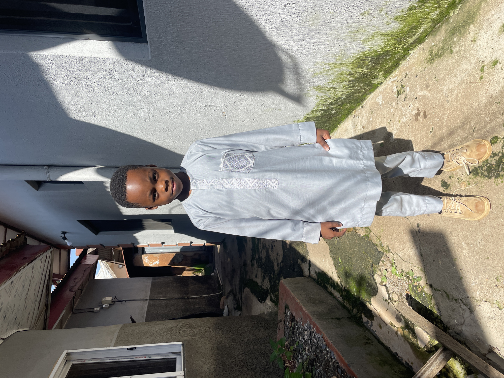A young boy stands in a narrow alleyway between two buildings in Jos, Nigeria. He is wearing a light blue traditional outfit with intricate white embroidery on the chest and a pocket. The boy has short black hair and is looking directly at the camera with a neutral expression. His right hand is in his pocket, and he is wearing light brown lace-up boots.
The alleyway is paved with concrete, which shows cracks and has patches of green moss growing on it. To the left, there is a raised concrete planter filled with gravel and a few small green plants. The buildings on either side are made of concrete and painted white. Sunlight casts strong shadows across the scene, creating a stark contrast between light and dark areas. The building on the right has a textured white wall with a window and what appears to be a downspout. The building on the left has a window with a white frame and a darker section of the wall below it. The alleyway recedes into the background, where more buildings and structures are visible, suggesting an urban environment. The overall scene depicts a bright, sunny day in a residential or commercial area.