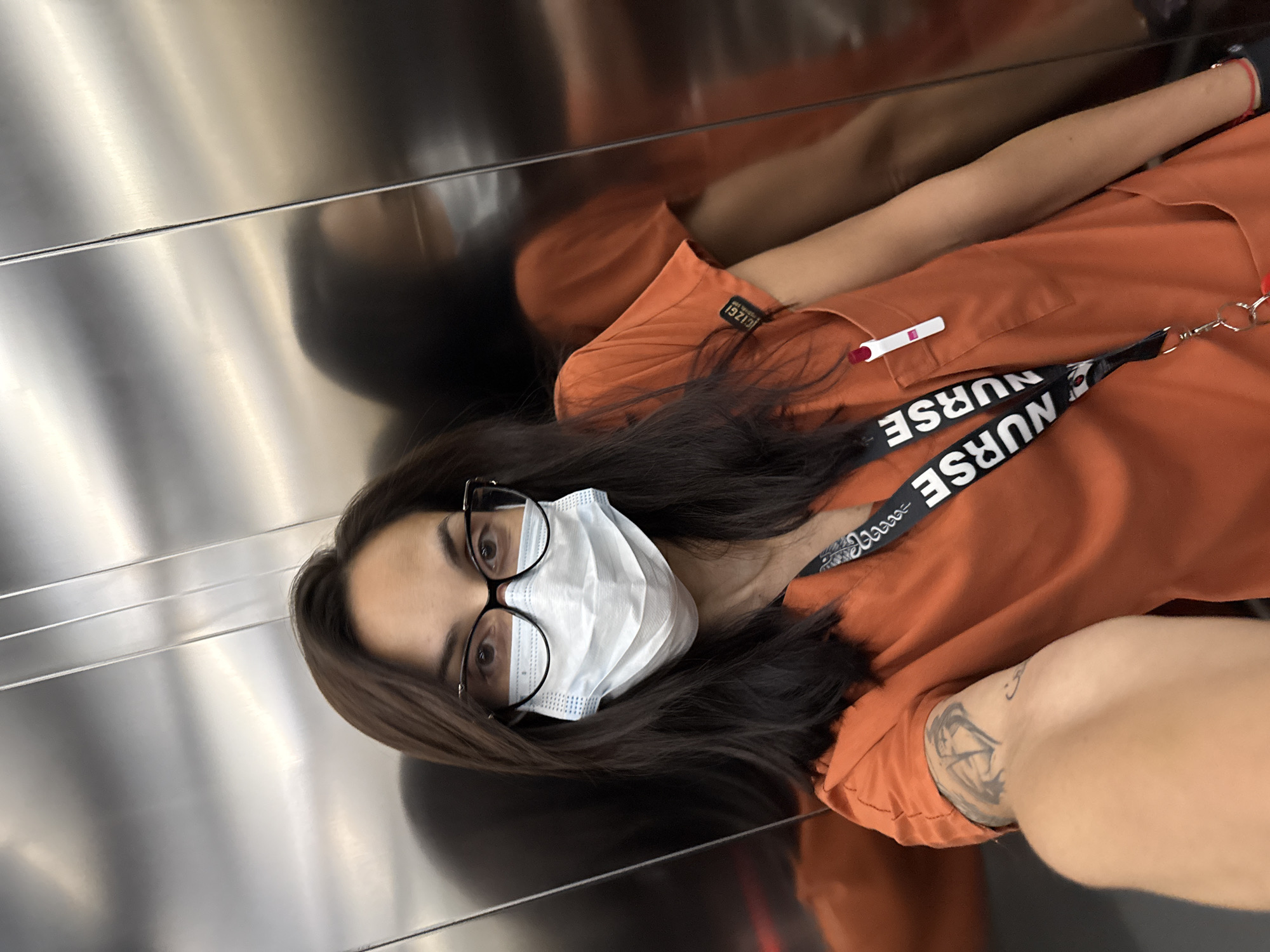 A woman with dark hair and wearing glasses and a white face mask is in an elevator. She's wearing an orange uniform with a "NURSE" lanyard and a pregnancy test clipped to her pocket. A tattoo is visible on her left forearm. The elevator has a polished metal finish. There is no information about Pleven, Bulgaria in the image.