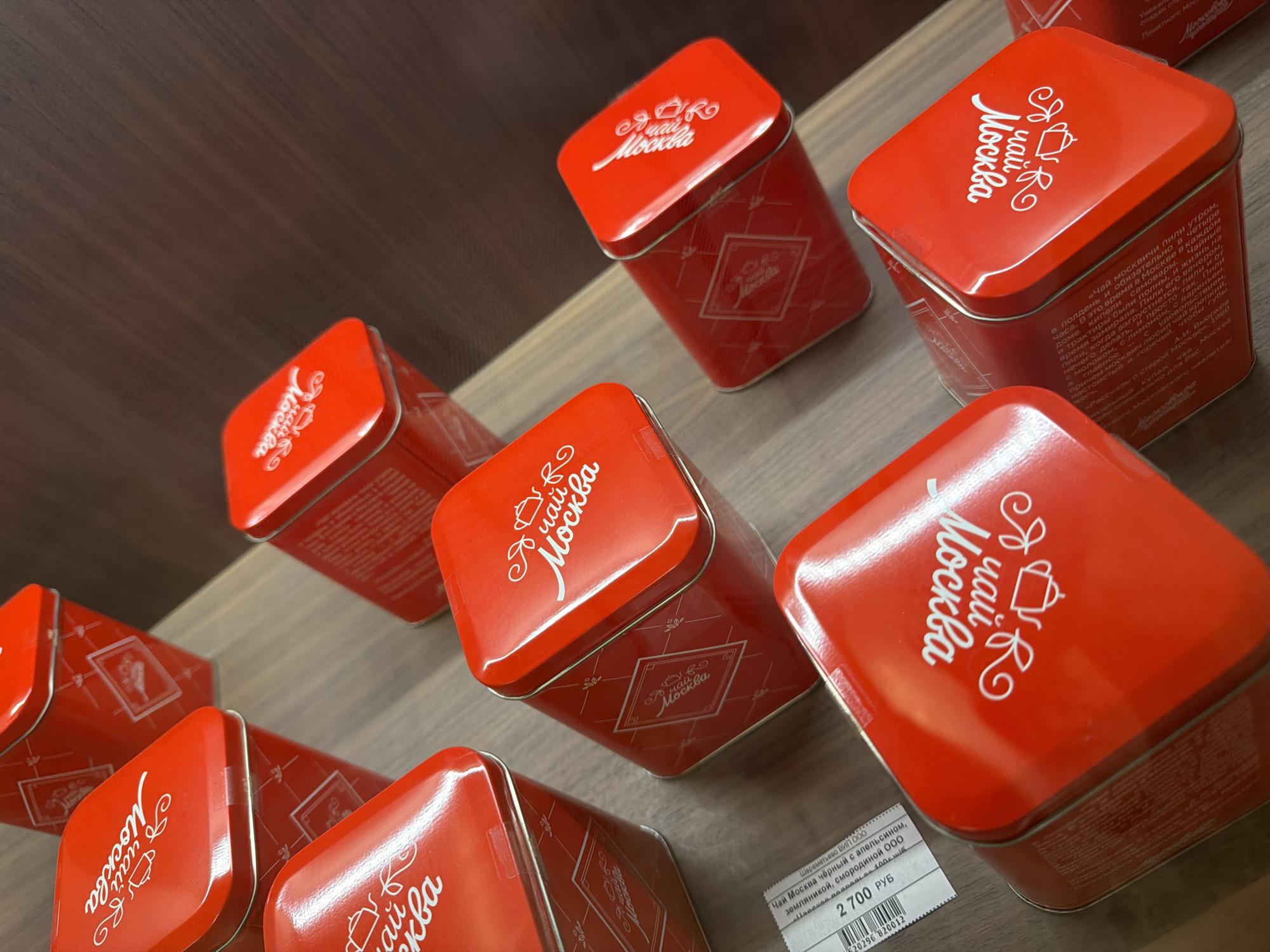 The image displays an angled, close-up view of several red tin containers, likely for tea, arranged on a wooden surface. The containers have white text and graphics, with the word "Москва" (Moscow) prominently featured on each. One visible label includes the text "Чай Московский черный с апельсином" (Moscow black tea with orange) and a price of "2 700 руб". The overall impression is of a retail display. The scene is indoors, with the wooden surface suggesting a shelf or counter. There are no people or animals visible in the image.