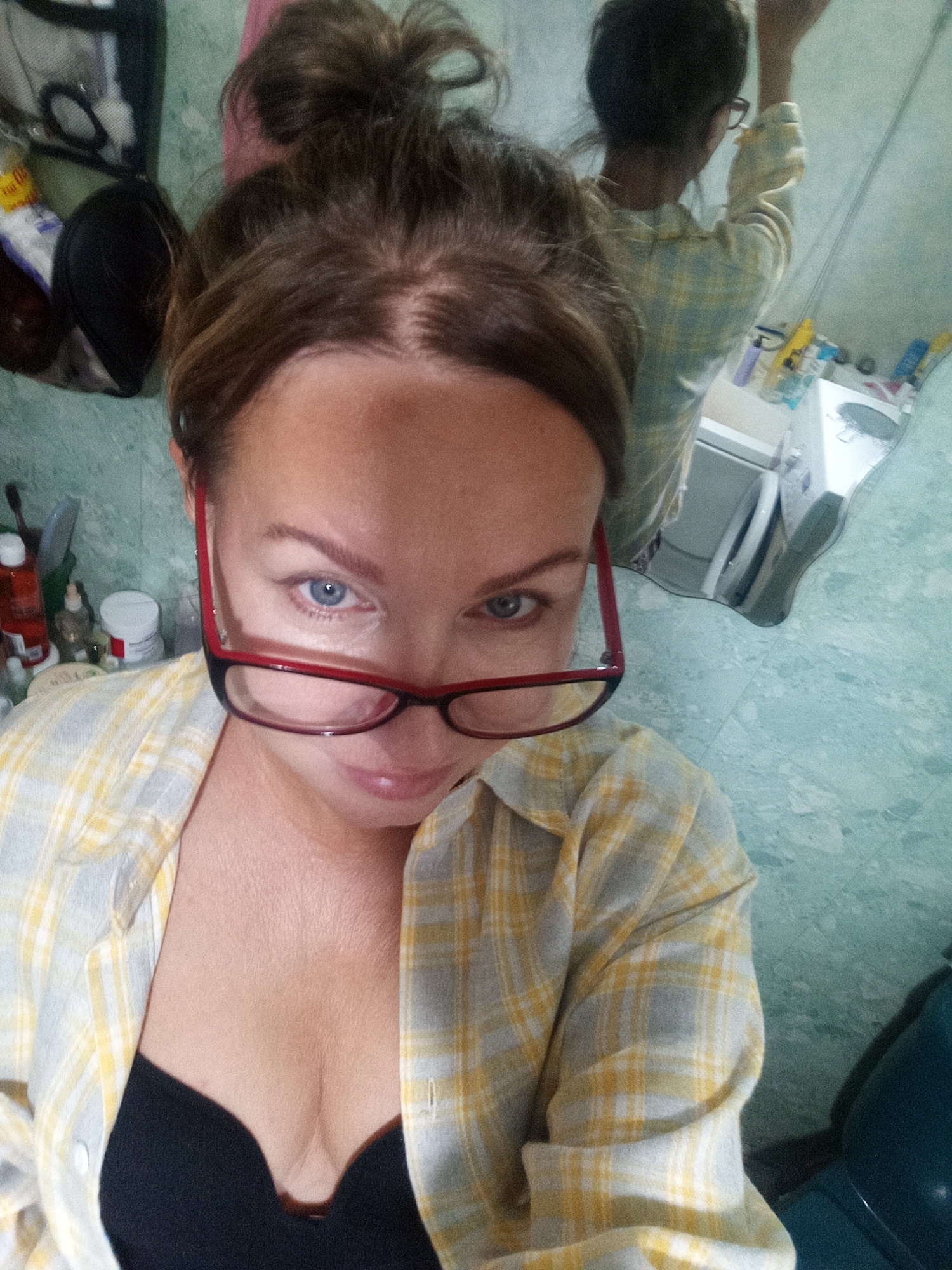 A woman with brown hair in a bun, wearing a yellow and white plaid shirt and a black bra, looks into the camera. She is wearing red-rimmed glasses. The background appears to be a bathroom with green tiled walls, a mirror reflecting another person, and various toiletries on a shelf.