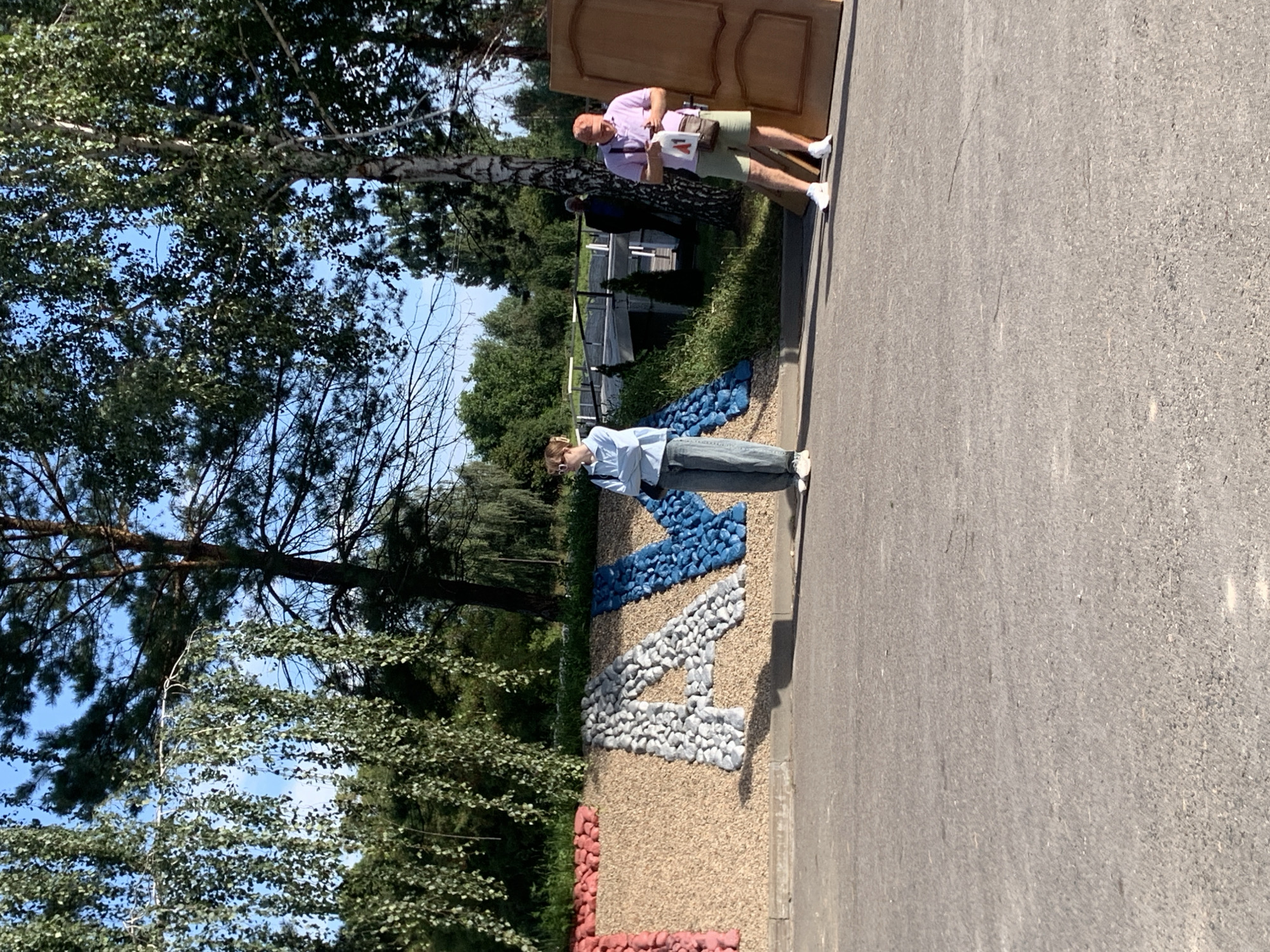 In a sunlit outdoor scene, a large, textured sign spelling out "FAM" with stones in red, grey, and blue is displayed against a wall covered in light brown gravel. To the right of the sign, a young person stands with arms crossed, wearing a light blue hoodie and grey pants. Further to the right, a man in a pink polo shirt and khaki shorts stands near a large, light brown wooden door, holding a phone and a bag. The background features lush green trees, a bright blue sky, and a bridge structure. The ground in the foreground is paved asphalt. The scene appears to be in the Бокшыцкі сельскі Савет, Belarus, based on the context provided.