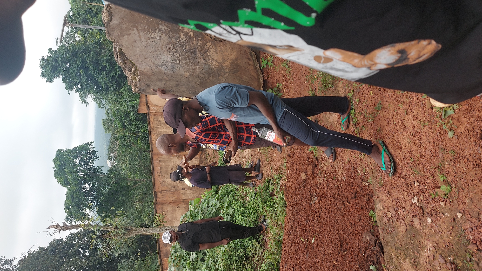 A group of five men are gathered outdoors in what appears to be a semi-rural area of Igbere, Nigeria. The ground is a mix of reddish-brown soil, gravel, and sparse vegetation. Behind the men, a weathered, pale orange wall stretches horizontally, with dense green foliage visible above it. The sky is overcast.
In the foreground, on the right, a man wearing a black t-shirt with a graphic print and dark pants is partially visible, facing away from the camera. He is wearing flip-flops. To his left, a man wearing a blue t-shirt and dark pants with a white stripe down the side is holding a bottle of "LORD'S" brand liquor. He is also wearing flip-flops. He is facing to his right, appearing to interact with the man next to him.
The man to the left of the liquor-holder is wearing a red and blue checkered long-sleeved shirt and dark pants. He is holding a dark object, possibly a phone, and is gesturing with his right hand, looking towards the man in the blue shirt. He is also wearing flip-flops.
Further to the left, a man in a black t-shirt with a silver and black design and black pants stands facing the camera. He is wearing a checkered cap. He is standing near a pile of reddish-brown dirt and lush green plants.
In the background, another man wearing a dark t-shirt and shorts is visible. He is wearing a black baseball cap and dark flip-flops.
The overall scene suggests a casual gathering outdoors, with the presence of alcohol indicating a social event. The location appears to be somewhat undeveloped, with a mix of natural elements and rudimentary structures.