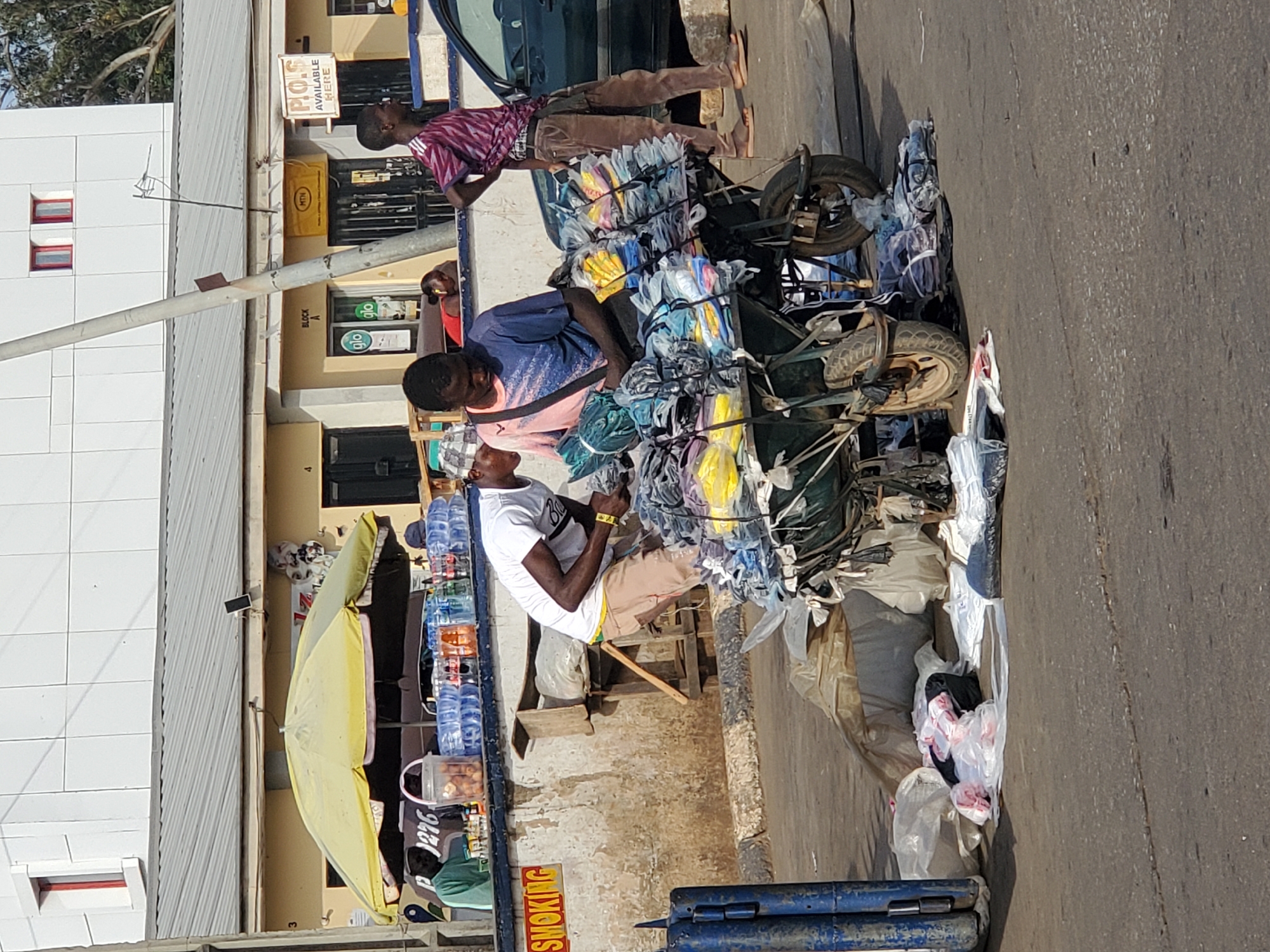 This image captures a vibrant street scene in Jos, Nigeria, under bright daylight, suggesting mid-morning to early afternoon. The setting is a commercial street bordered by buildings, including a modern white structure and an older beige building with multiple businesses.
Several men are visible, engaged in street vending activities. In the center, a man in a blue and pink shirt is seated on a heavily laden three-wheeled motorized cart, packed with numerous bundles of what appear to be textiles or clothing, individually wrapped in clear plastic. Another man in a white t-shirt and light brown shorts sits on a wooden stool facing him, possibly a customer or another vendor. Behind them, a man in a striped shirt and brown trousers stands next to a dark vehicle, observing the scene.
To the left, a yellow market umbrella provides shade over a smaller stall, displaying bottled water and other items, with another person partially visible behind it. The street itself is paved, with some debris and bags on the side, typical of an active urban market. In the foreground, blue metal barriers are partially visible.
Visible text on the buildings provides location cues: a sign reads "P.O.S AVAILABLE HERE," and below it, a yellow sign bears the "MTN" logo. Further down the building, the "glo glo" telecom logos are present, along with "BLOCK A." A partially obscured red sign near the bottom right reads "NO SMOKING." The overall atmosphere is busy and commercial, characteristic of street trade in a Nigerian city.