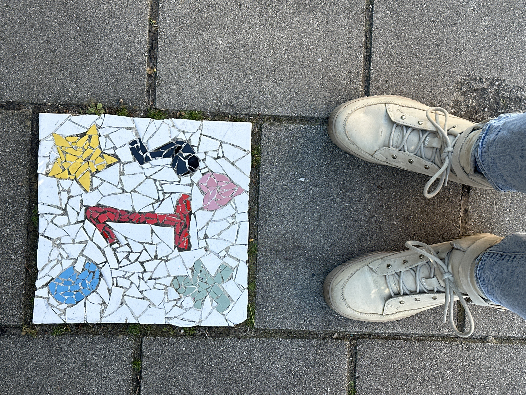 A mosaic tile of broken pieces with a yellow star, black shape, pink heart, red angular figure, blue blob, and green cross is set into a gray concrete walkway, flanked by two feet in light-colored shoes and blue jeans. The location is in Amsterdam, Netherlands, based on the known mosaic patterns found in its public walkways.