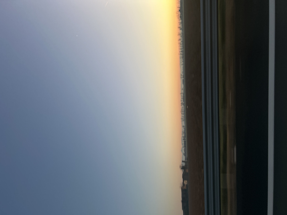 The image, when correctly oriented to landscape, presents a serene view of a vast sky and horizon, captured during either sunrise or sunset. The upper half of the scene is dominated by a clear sky displaying a striking color gradient; it transitions from a soft, deep blue on the left to a vibrant, warm orange-yellow on the right, indicating the low position of the sun. A faint, thin white line, possibly an airplane contrail, is visible in the upper left portion of the blue sky.
A dark, indistinct horizon line stretches horizontally across the middle of the image, separating the colorful sky from the ground below. This distant line suggests a sparse landscape, likely featuring very small, unidentifiable structures or trees silhouetted against the bright sky. The immediate foreground below this horizon is dark and lacks specific detail, appearing as a flat expanse of land.
On the far right side of the image, a prominent, dark vertical structure partially frames the scene. This element strongly suggests the photograph was taken from an interior viewpoint, such as through a window of a building or a moving vehicle. This structure features several thin, parallel horizontal lines, possibly indicating a textured surface or functional elements of the frame. There are no people or visible text in the image. The overall impression is one of a peaceful and expansive natural vista during the golden hour.