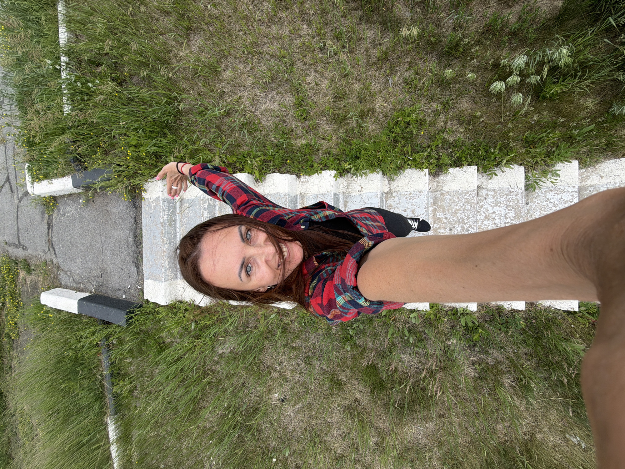 A young woman is captured from a high-angle perspective, looking up at the camera with a wide smile. She is wearing a red and blue plaid shirt over a black top, black pants, and black and white sneakers. Her long brown hair is partially obscuring her face. The woman is standing on a set of outdoor concrete stairs that are bordered by patches of grass and small yellow wildflowers. To the left of the stairs, there is a concrete surface and a black and white marker post. The scene appears to be outdoors during the daytime, with overcast skies suggested by the lighting. The overall impression is one of a casual, candid moment, possibly a selfie, with the woman expressing joy and engagement with the camera. The location is specified as Nikolaevka, Russia.