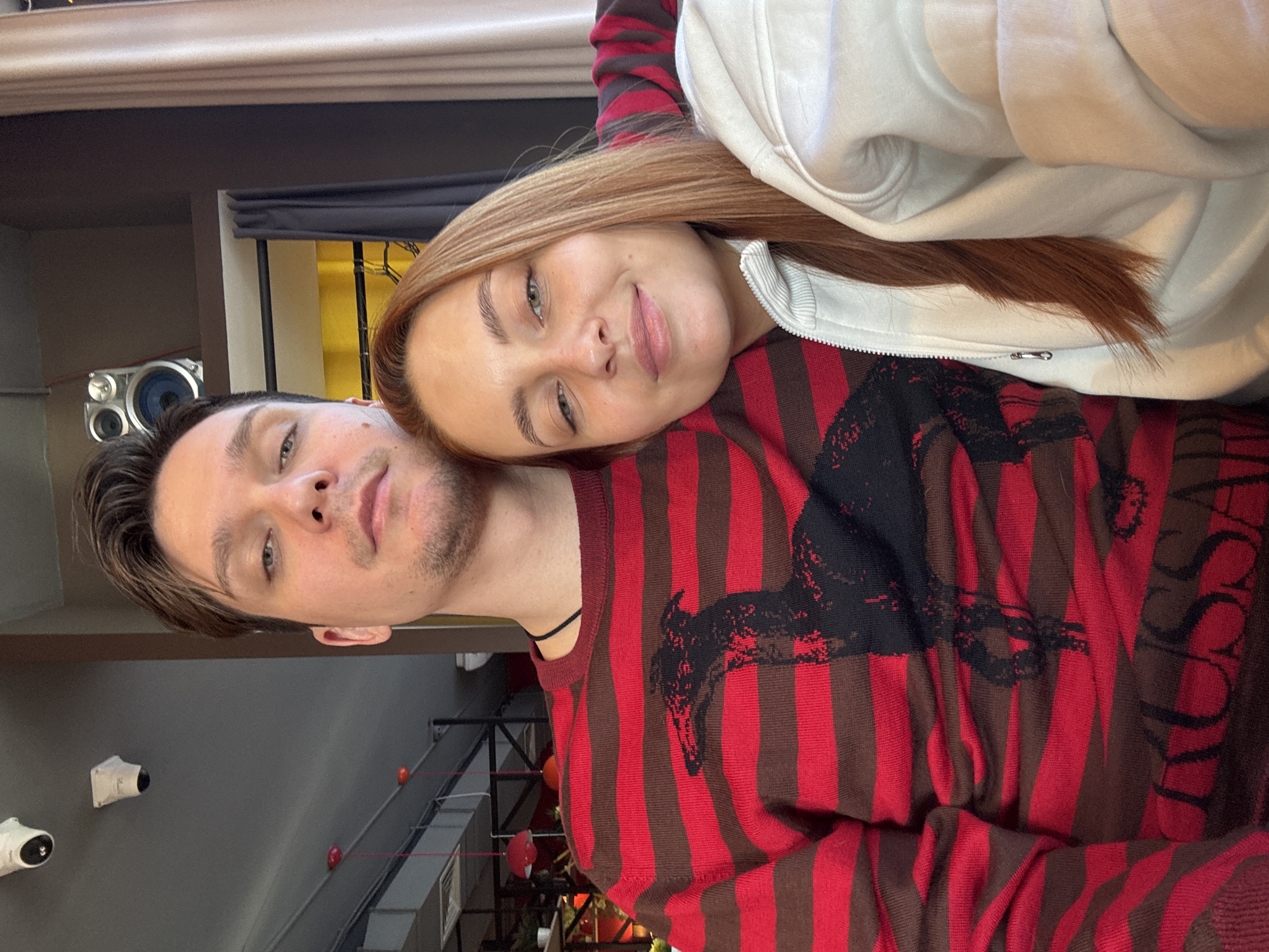 A selfie taken indoors shows a man and a woman. The woman rests her head on the man's shoulder. The man is wearing a red and brown striped sweater with a black design. The woman is wearing a white hoodie. The background includes a speaker, curtains, and what appears to be a wall with security cameras. Based on the location provided, this photograph was taken in the city of Chaikovsky, Russia.