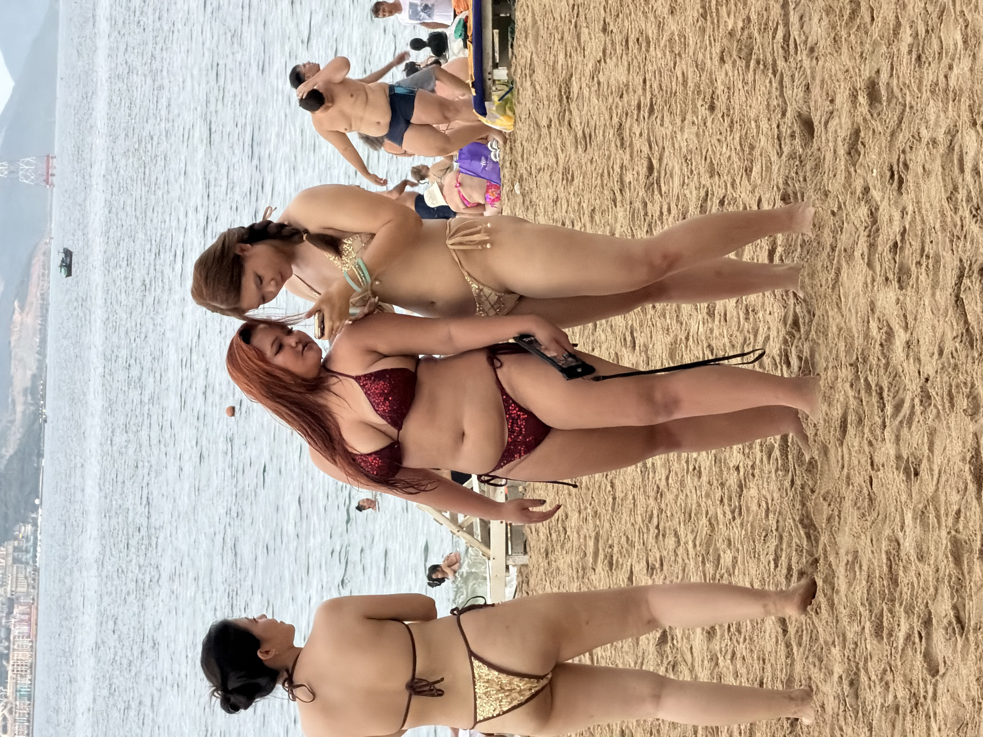 The image depicts a crowded beach scene in Nha Trang, Vietnam. In the foreground, three women are the primary subjects. On the left, a woman with her back to the camera wears a dark string bikini with a gold sequined bottom. The two women in the center are facing each other and looking at a smartphone. The woman on the right has long braided hair and wears a gold sequined bikini. The woman to her left has red hair and is wearing a dark red sequined bikini. The beach is sandy, with the ocean visible in the background. Other beachgoers are scattered across the sand and in the water. In the distance, buildings and a tower are visible on the coastline, characteristic of Nha Trang's urban beach landscape. The water is a dull blue-grey, suggesting overcast weather or choppy conditions.