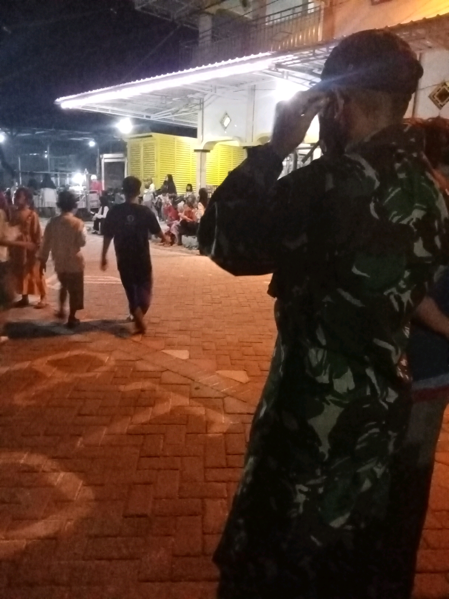 The image depicts a nighttime scene on a brick-paved walkway in Keplaksari, Indonesia. In the foreground, a person wearing a camouflage uniform and a dark cap salutes, obscuring their face. Behind them, several people are visible, some walking and others seated. Two young individuals are walking away from the viewer, one in a dark t-shirt and the other in a lighter shirt. A brightly lit yellow structure with an awning is visible in the background, with more people gathered around it. The overall atmosphere suggests a public gathering or event at night.