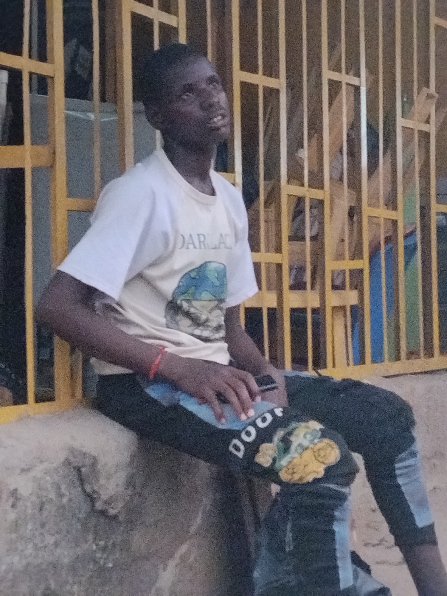 The photograph shows a young man in Ura Nta Umuarandu, Nigeria, sitting on a ledge with a yellow metal gate behind him. He is wearing a white t-shirt with a graphic and the word "DARLA" printed on it, along with dark pants that have light blue accents and an embroidered design on the knee. He is looking upwards and slightly to the right. His left hand is resting on his lap. The surrounding environment consists of what appears to be a partially blurred building exterior.