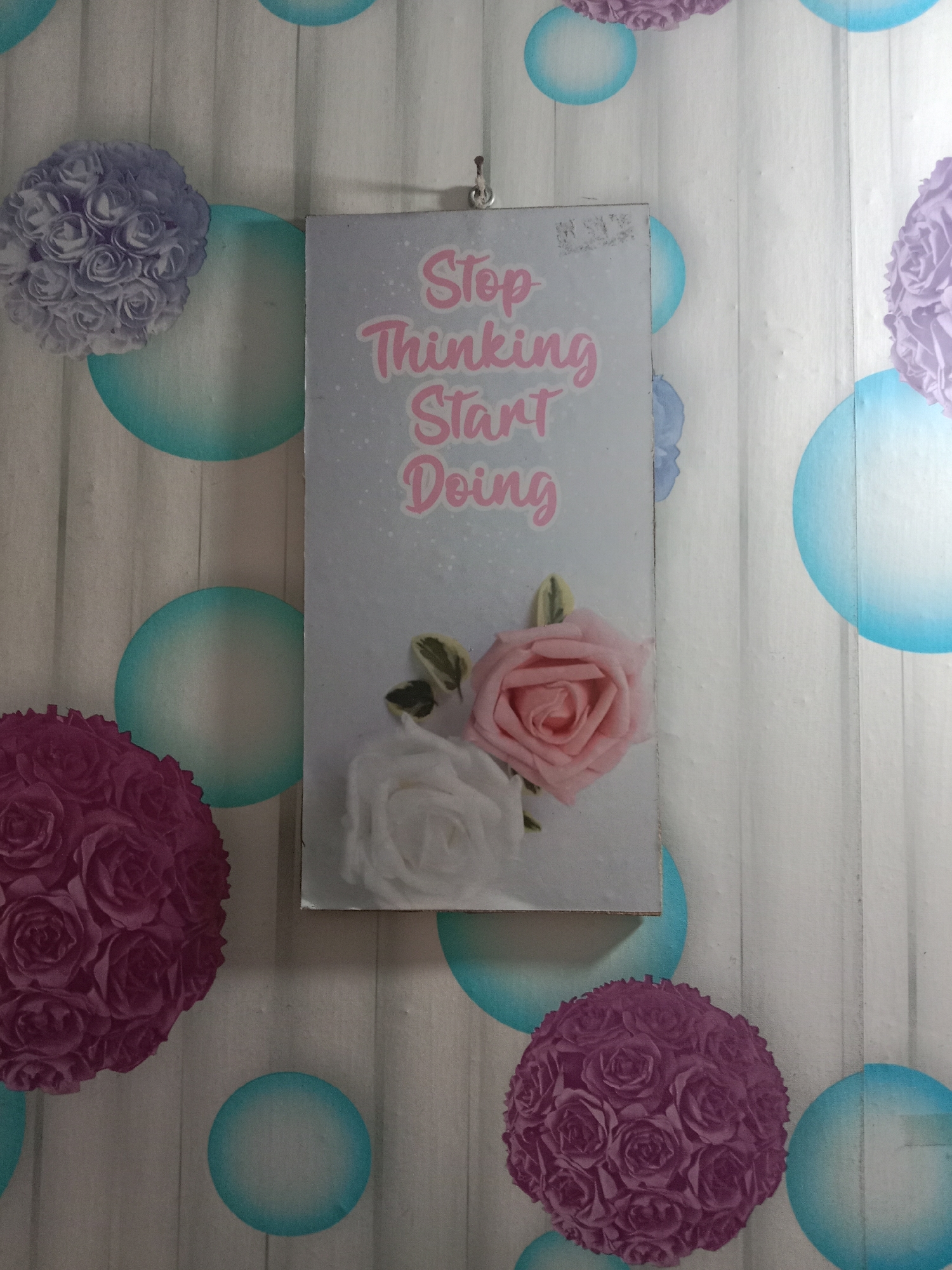 A framed inspirational quote with an image of roses is hanging on a wall. The quote reads "Stop Thinking Start Doing" in pink script. The wall is decorated with a pattern of light grey wood planks and various sizes of teal and light blue circles that resemble bubbles or balloons. There are also floral decorations on the wall, including a large cluster of purple roses on the left side and another cluster of purple roses on the bottom right. A pink rose and a white rose are depicted on the framed quote itself, as well as some green leaves. The scene is indoors and appears to be in a residential or commercial space, possibly in the city of Kutosari, Indonesia, based on the provided information.