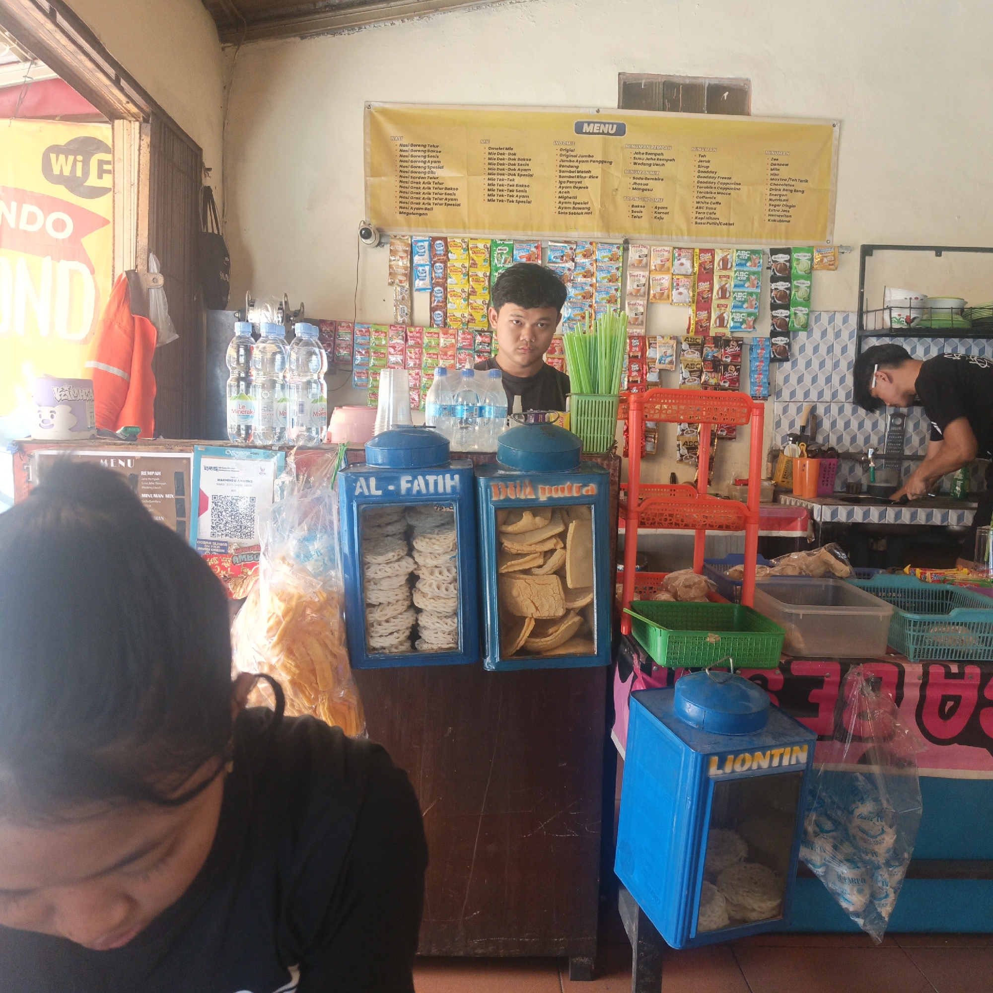 The image depicts a small food stall, likely a warung or kedai, operating in Semarang, Indonesia. The stall is characterized by its rustic setup, with a counter displaying various food items and beverages. Behind the counter, a young man, presumably the vendor, is visible. He is wearing a black t-shirt and appears to be interacting with a customer or attending to the stall's operations.
The stall is stocked with an assortment of goods, including bottled water, packets of snacks, and what appear to be freshly prepared snacks in transparent containers. A menu is displayed prominently on a yellow banner, listing various food and drink options, including "Nasi Goreng Telur" (Egg Fried Rice), "Mie Goreng Sosis" (Sausage Fried Noodles), and various types of beverages such as "Teh" (Tea), "Kopi" (Coffee), and "Jus" (Juice). The presence of a QR code suggests that cashless payment options might be available.
The overall environment is that of a typical street-side eatery, with basic furnishings and a focus on serving local Indonesian cuisine and refreshments. The visual cues, such as the menu items and the general ambiance, strongly suggest a location within Semarang, given the prevalence of such establishments in the city.
