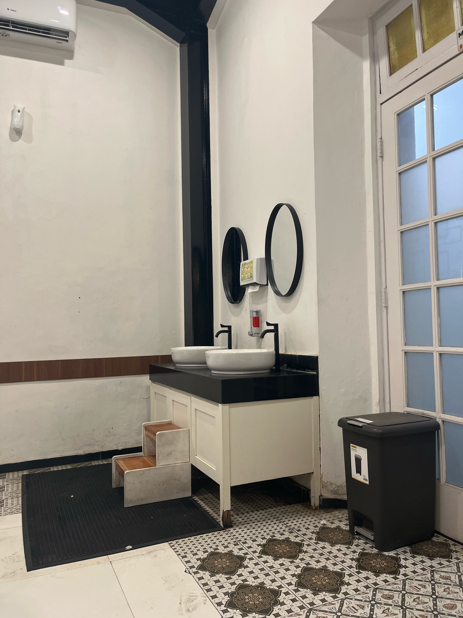 The image shows a bathroom area with two sinks mounted on a white vanity. Above the sinks are two oval mirrors and a dispenser. To the left of the vanity is a black mat with a small set of wooden steps. To the right of the vanity is a black trash can with a label. The floor is tiled with a patterned design. A white door with glass panes is visible on the right side of the image. An air conditioning unit is mounted high on the wall to the left. The scene appears to be a public restroom, possibly in a commercial establishment. This location is identified as a restroom in Semarang, Indonesia.