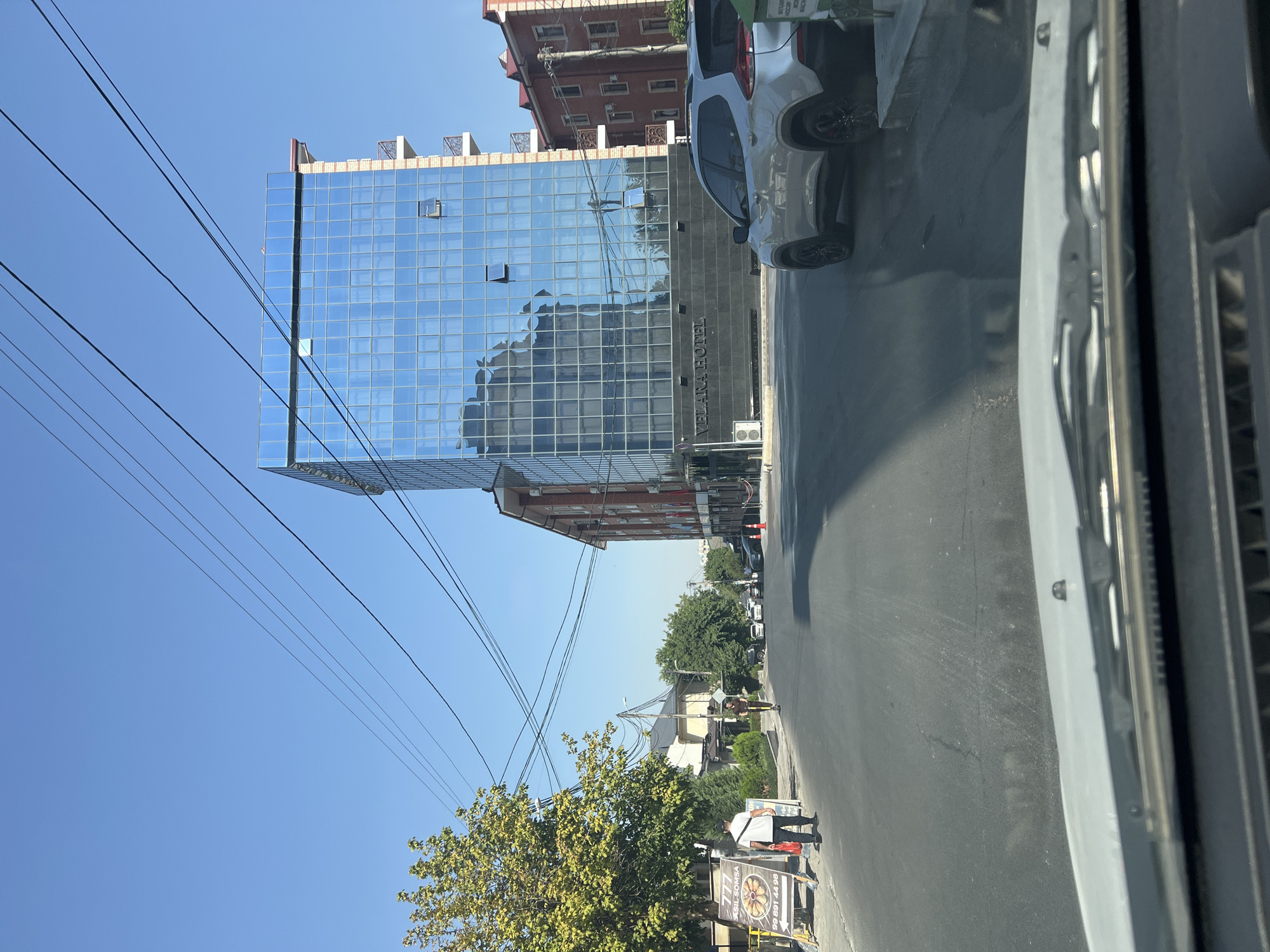 The image shows a street scene in Tashkent, Uzbekistan, under a clear blue sky. A modern glass-fronted building, identified by the text "VELARA HOTEL" on its facade, dominates the right side of the frame. Adjacent to it is a brick building with balconies. A silver SUV is parked on the right side of the street. On the left side of the street, a man in a white shirt and dark pants is walking away from the camera, carrying a red bag and a white object. In front of him is a stand with advertisements. Further down the street, a person is riding an electric scooter. The road is paved and appears to be a city street with power lines overhead. The environment suggests a sunny day in a developed urban area.