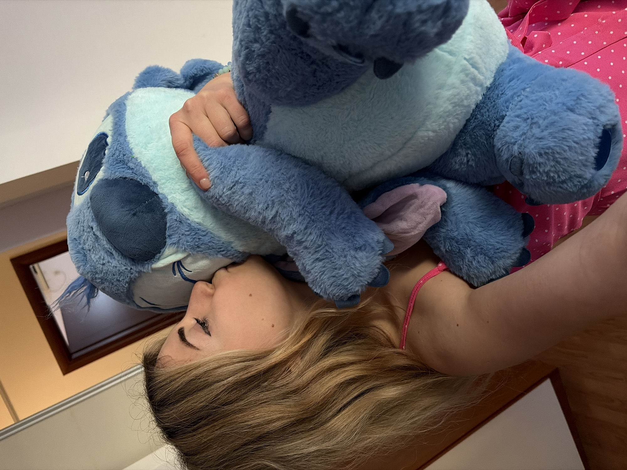 A woman is lying down and cuddling a large, blue plush toy of the character Stitch from the movie "Lilo & Stitch". She is holding the toy with one hand. The woman has blonde hair, fair skin, and is wearing a pink top. She is looking towards the toy and appears to be kissing it. The background is a room with a wooden floor and a picture frame on the wall. There is no information in the image that reveals if it was taken in Budva, Montenegro.