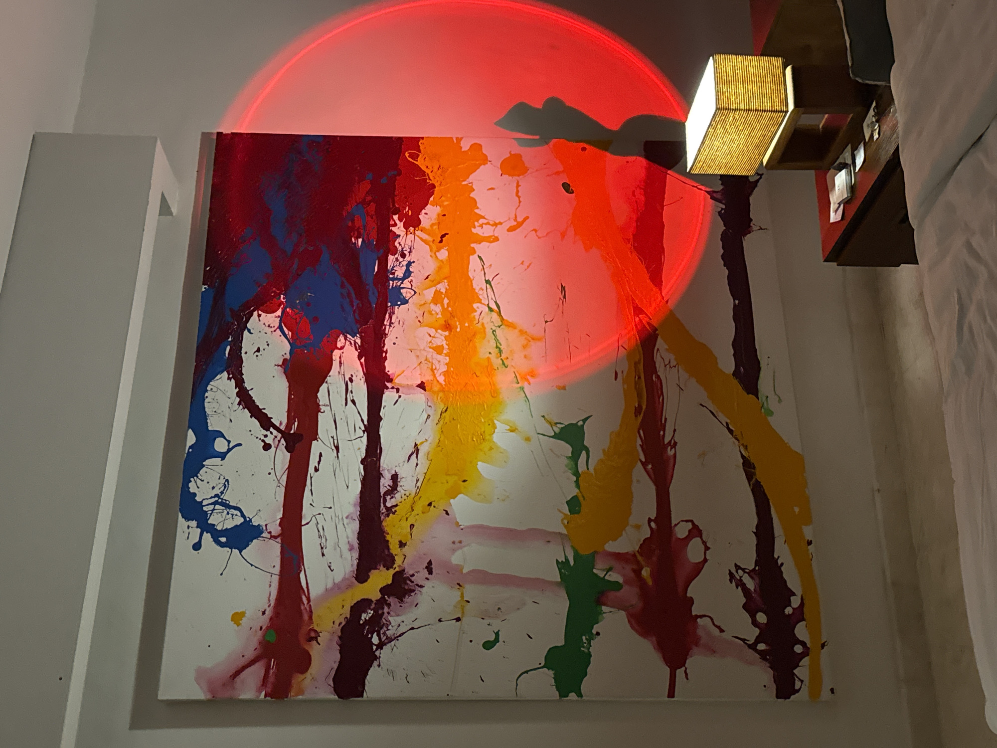 The image displays an artistic indoor setting, likely a bedroom or living space in North Kuta, Indonesia. Central to the frame is a large, abstract splatter painting on a white canvas, characterized by vibrant drips and splashes of red, blue, orange, yellow, and green paint.
A prominent feature is a circular red light projected onto the wall, overlapping the top section of the painting and casting an atmospheric glow. In the upper right corner, a modern, rectangular lamp with a slatted wooden shade emits a warm, yellowish light, adding to the room's illumination. Below this lamp, a wooden headboard or shelf is partially visible, alongside a white bedsheet, suggesting a sleeping area. To the left, a white architectural element, possibly a column or corner of a wall, frames the scene.
The lighting conditions indicate it is evening or night. No people are visible, nor is there any discernible text within the image. The arrangement of the colorful artwork and the distinct lighting creates a dynamic and artistic ambiance within this interior space.