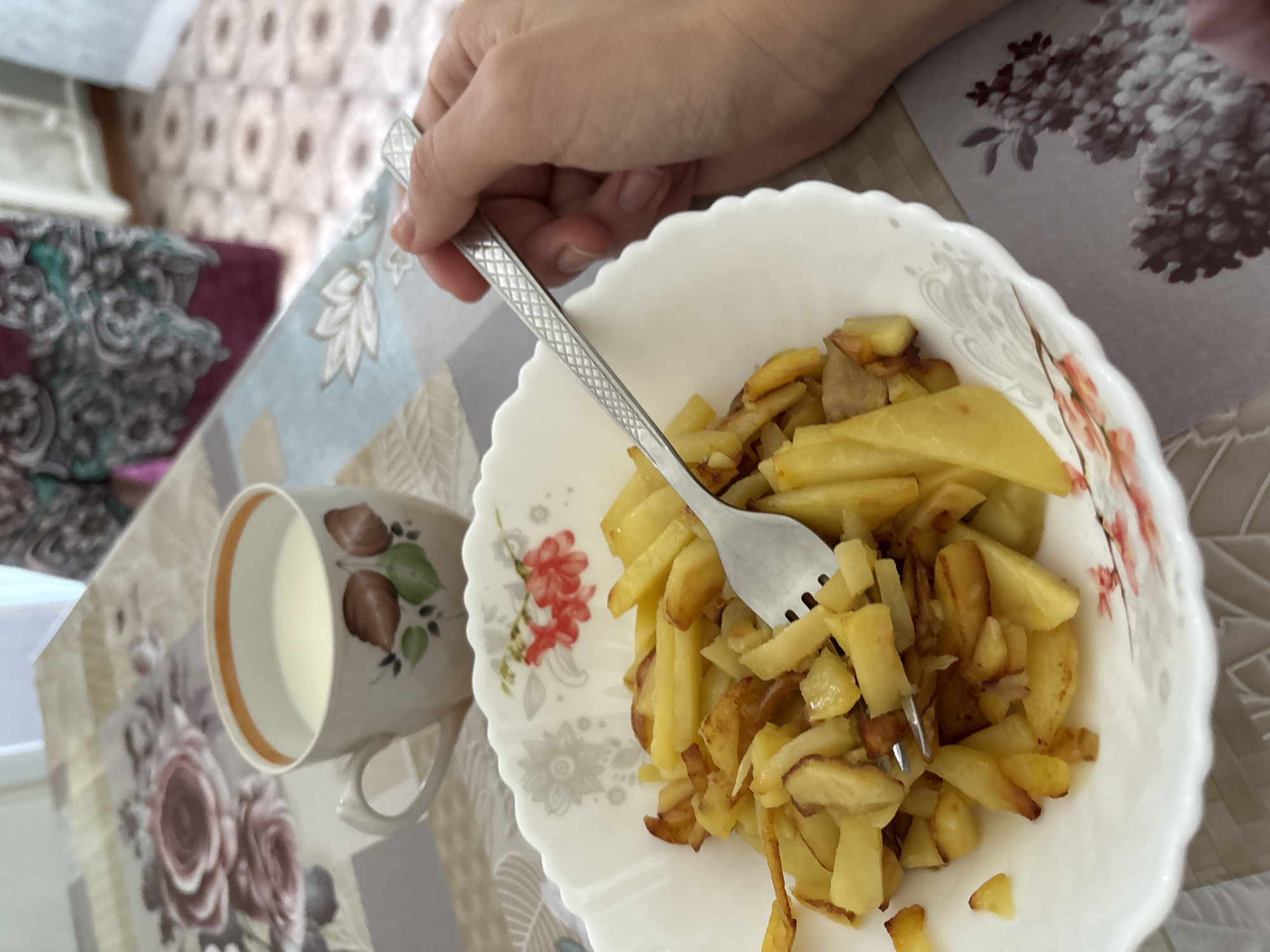 A person's hand holds a fork, poised over a bowl of fried potatoes. A mug of milk is positioned to the left of the bowl. The table is covered with a floral tablecloth. The setting appears to be indoors. It is not possible to determine the location with certainty.