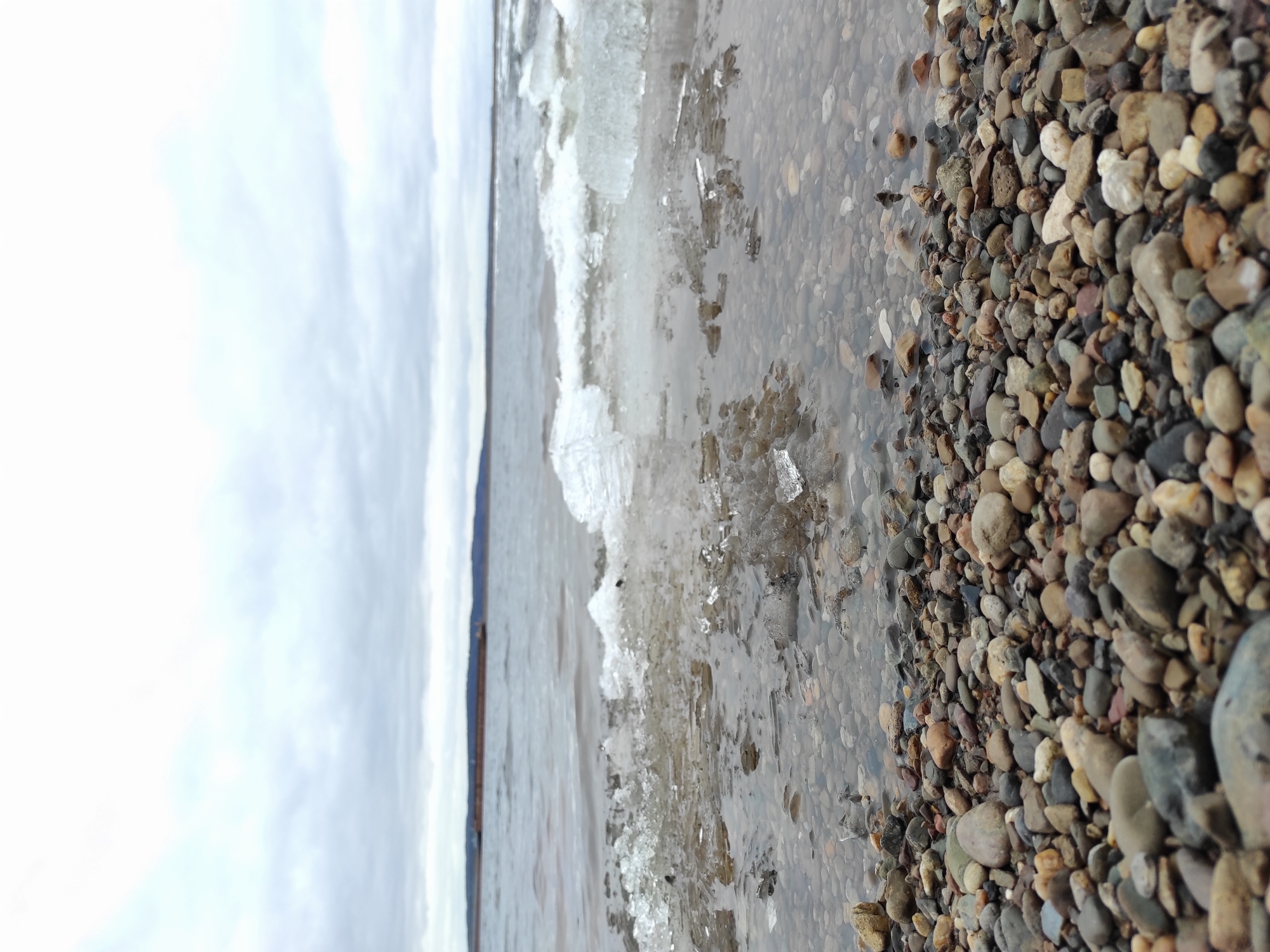 The image depicts a shoreline with a stony beach in what appears to be a body of water. The foreground shows a collection of multi-colored pebbles and small rocks, some partially submerged in water. Moving towards the center, there are pieces of ice scattered along the waterline, interspersed with shallow water. In the background, a strip of land is visible beneath a cloudy sky. The scene likely takes place on a cold day due to the presence of ice. The exact location within the city of Chaikovsky, Russia, cannot be determined from the visual information provided in the image. There are no people visible in the image.