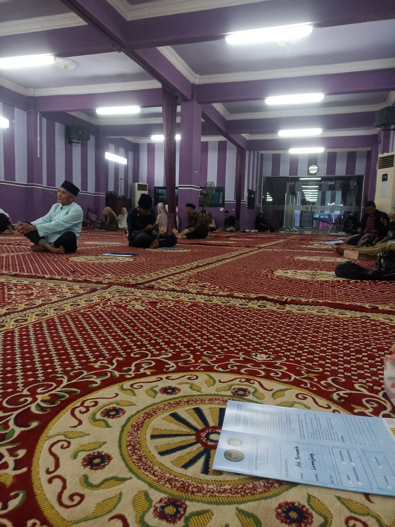 The image depicts the interior of a prayer hall, likely a mosque or similar place of worship, in Tompokersari, Indonesia. The hall is spacious with a high ceiling adorned with purple and white striped walls and white trim. Fluorescent lights illuminate the room. The floor is covered by a large, ornate red carpet with intricate geometric and floral patterns in red, yellow, green, and blue.
Several people are seated on the carpet, facing in various directions. In the foreground, to the left, a man wearing a light blue shirt and a black cap is seated with his legs crossed, looking to his left. To his right, another man in a dark shirt and black cap is also seated and appears to be looking at something in his hands. Further back, other individuals are seated on the carpet, some with head coverings.
In the foreground, a document, possibly a form or application, lies open on the carpet. It is a light blue and white paper with printed text and what appear to be boxes for filling in information. Two circular seals are visible on the document. The overall atmosphere suggests a gathering for prayer, study, or a community event within a religious setting. The image is taken from a low angle, emphasizing the expanse of the carpet and the ceiling.