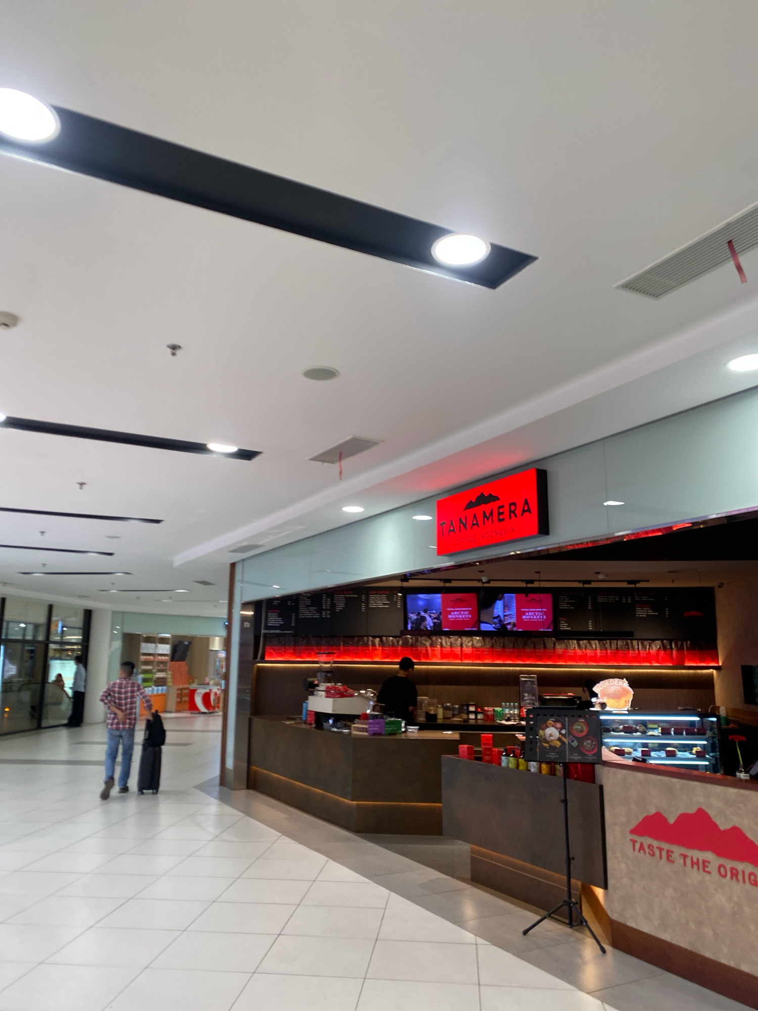 The image depicts the interior of a modern shopping mall in Batam, Indonesia, specifically focusing on a Tanamera Coffee shop. The coffee shop features a prominent red neon sign with the Tanamera logo and "COFFEE INDONESIA" written below. There are two digital menu screens displaying images, and the counter area is lined with various coffee-related items and red lighting. A person is seen working behind the counter, and another person is standing near a display case filled with pastries.
In the foreground, a man in a plaid shirt and jeans is walking away from the camera, pulling a black suitcase. He is on a tiled floor in a wide walkway of the mall. The ceiling of the mall has a contemporary design with recessed lighting and dark rectangular accents. Other parts of the mall are visible in the background, including shop fronts and shoppers. The overall atmosphere is bright and commercial.