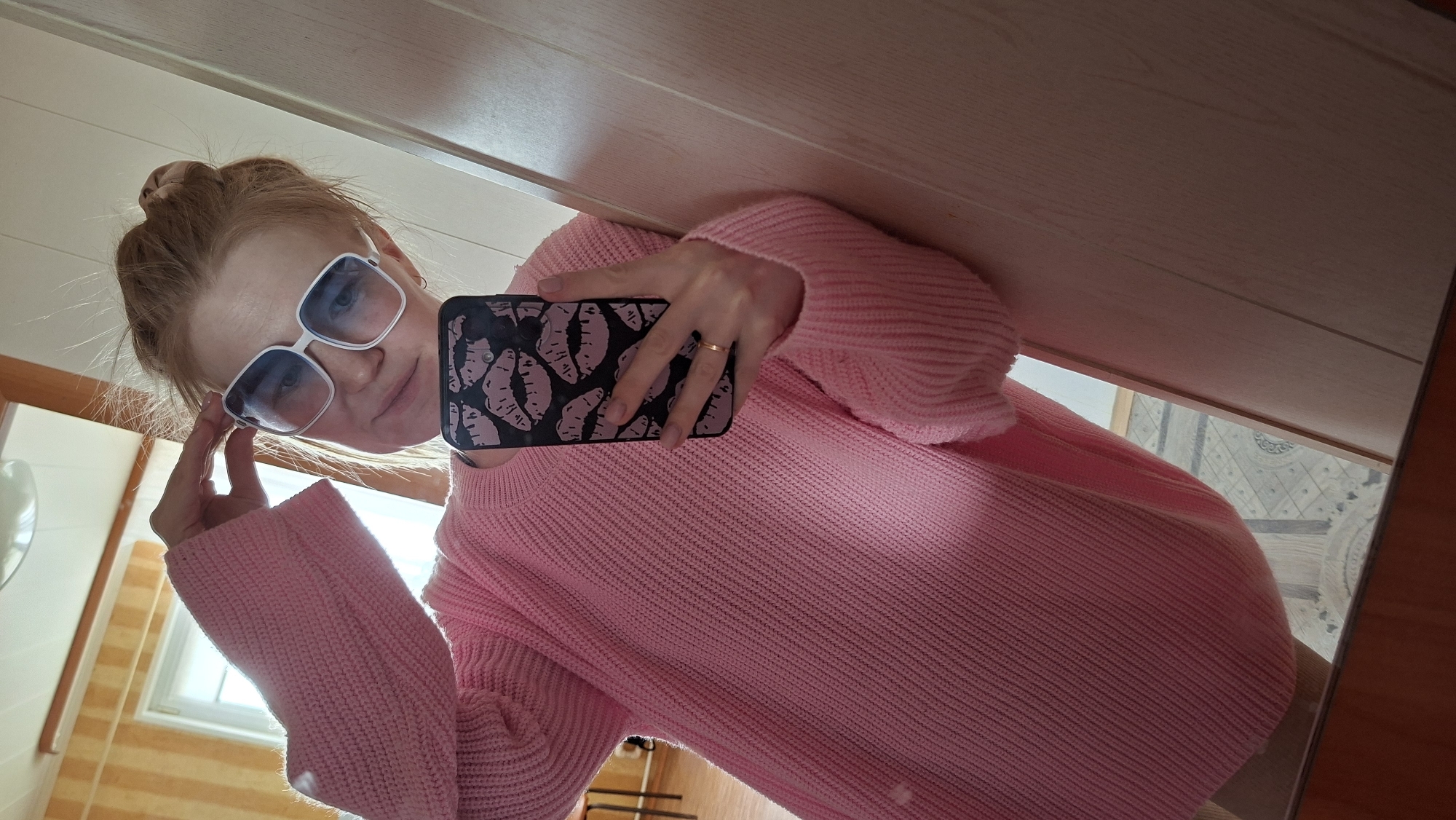 A woman in Большой Букор, Russia, poses for a selfie in a mirror. She is wearing a pink sweater and large, white-framed sunglasses with blue-tinted lenses. Her hair is pulled back into a bun. She is holding a black smartphone with a pink lips design on the case. The background appears to be a bathroom, with a window visible in the lower left and a door or cabinet above her.