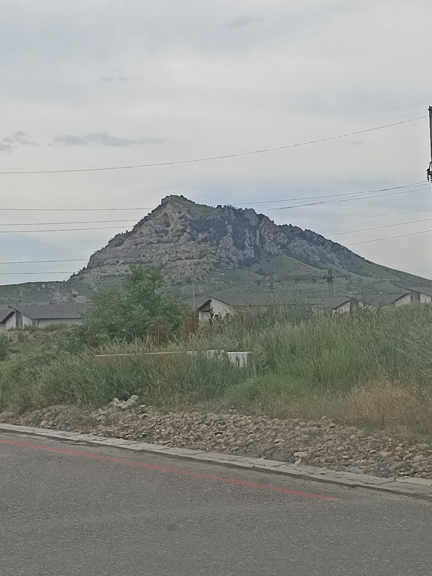 This is a medium shot of a mountain range, likely Vinsady, Russia, under a cloudy sky. The mountain in the background has a jagged, rocky surface and appears to be covered in sparse vegetation. There are several terraced roads or pathways visible on the mountain's slopes. In the middle ground, there is a cluster of buildings with dark roofs, some of which appear to be houses. A dense area of green grass and trees separates the buildings from the foreground. The foreground consists of a paved road with a red line marking its edge, and a rocky embankment. Power lines are visible in the sky, crossing horizontally across the frame. There are no people visible in the image.